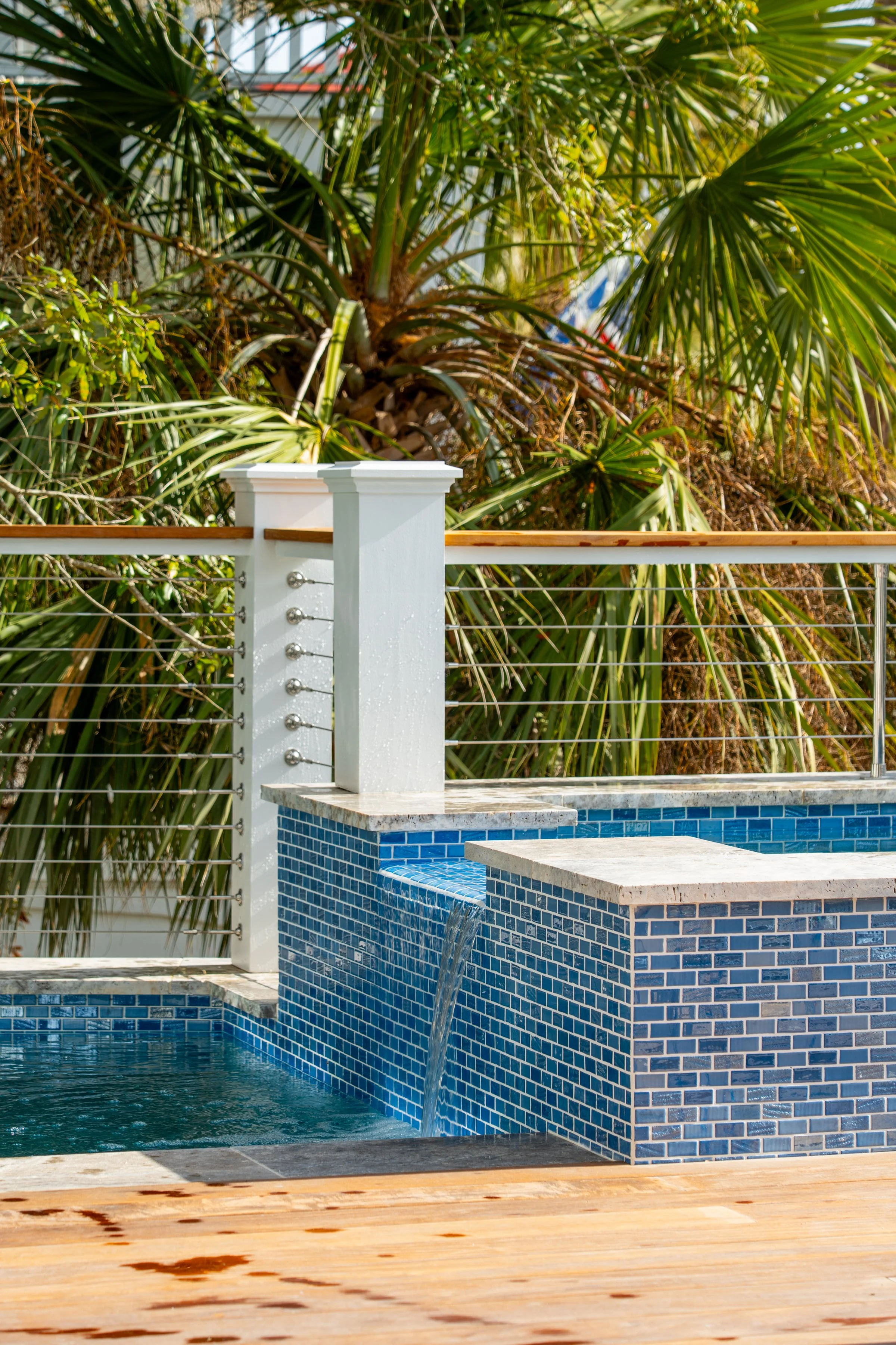 Close-up of a modern backyard pool with blue tile walls, a small waterfall feature, surrounded by a wooden deck and a white railing, with lush green palm trees in the background.
