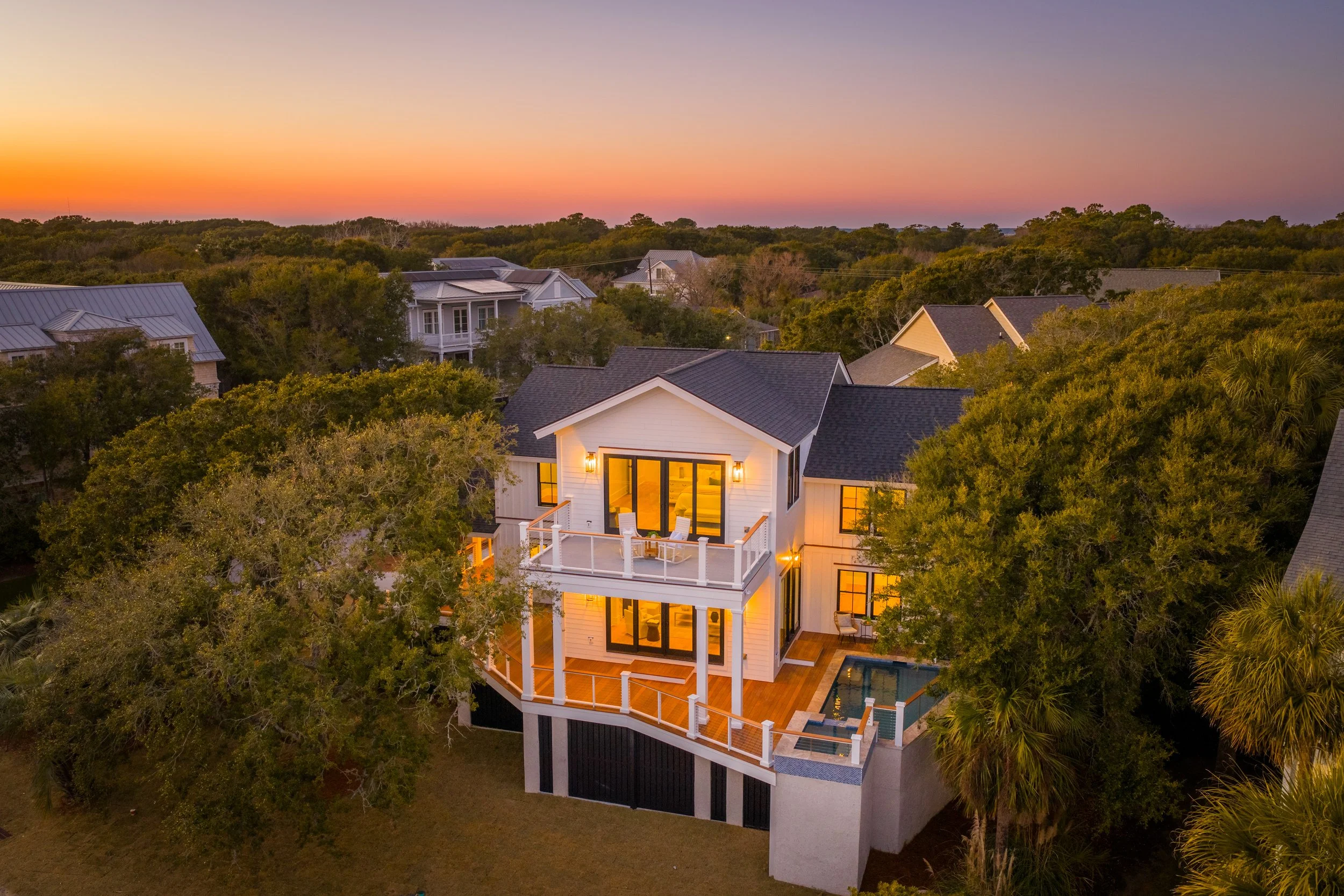 A modern house at sunset with illuminated windows and a pool on the deck, surrounded by trees and neighboring houses.