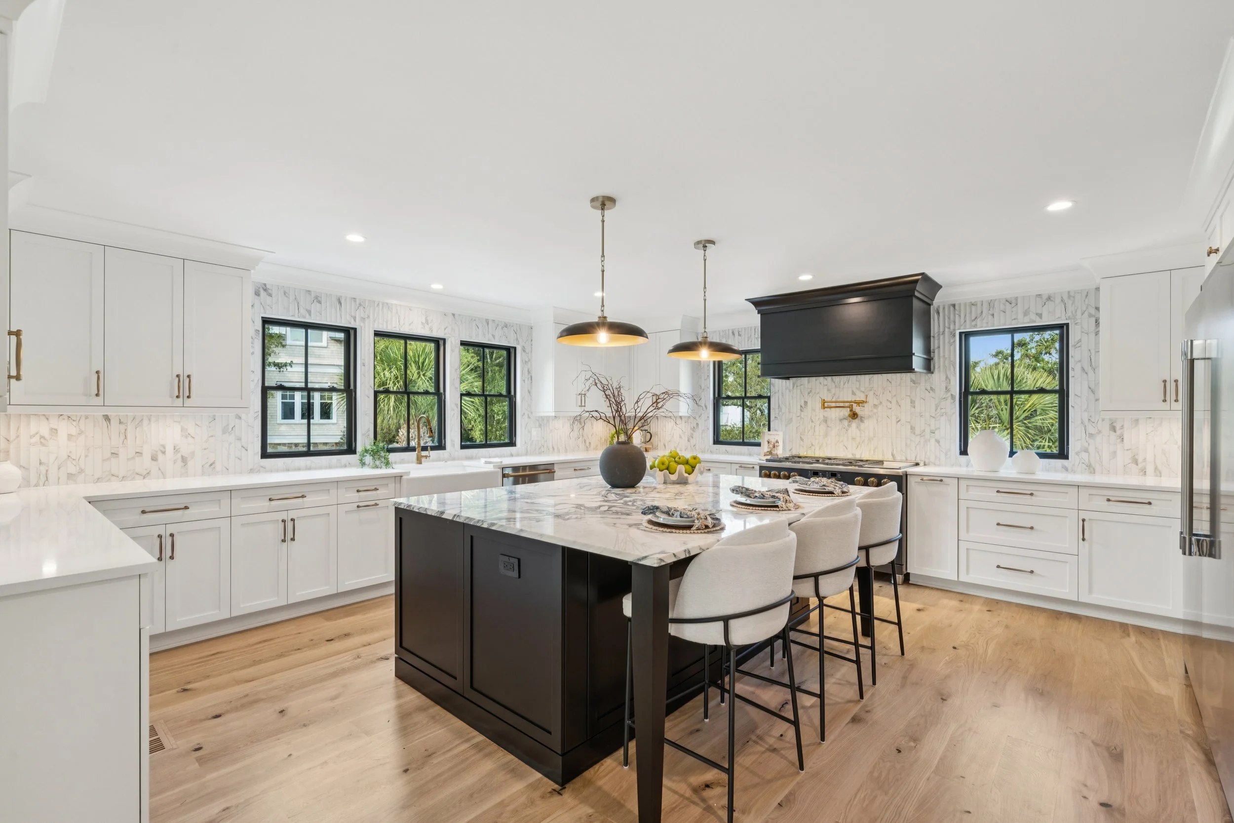 Bright modern kitchen with white cabinetry, black window frames, and a black island with marble countertop, four white bar stools, and pendant lights, large windows with greenery outside.