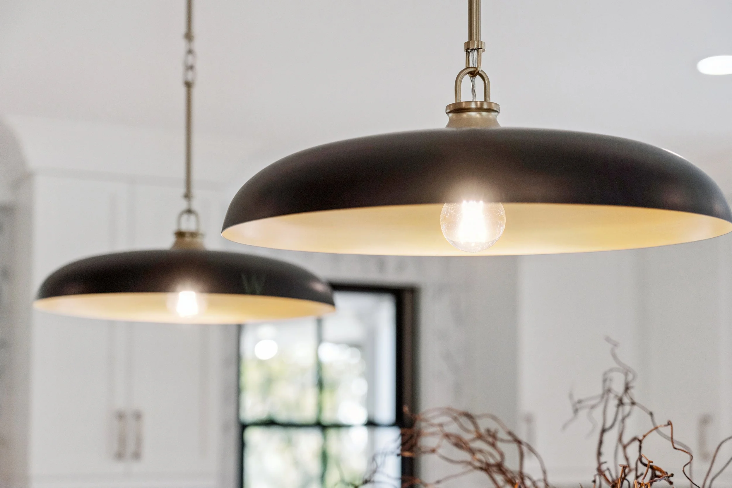 Two black pendant lights with brass accents hanging from ceiling in a modern kitchen, with a window and some decorative branches visible in the background.