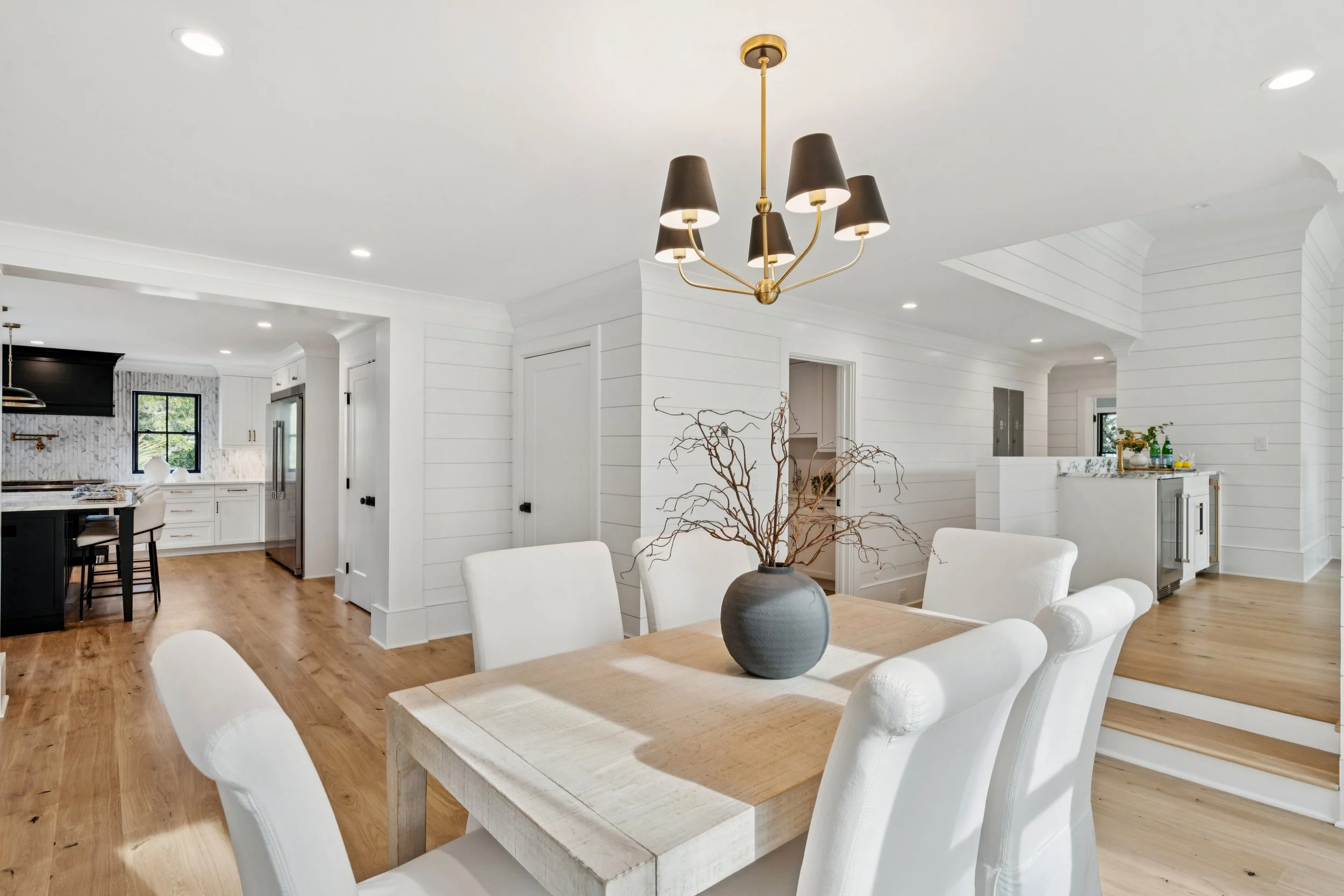 Modern open-plan dining area with a wooden table, white upholstered chairs, a black and gold chandelier, and a decorative vase with branches, connected to a kitchen with white cabinetry and black countertops.