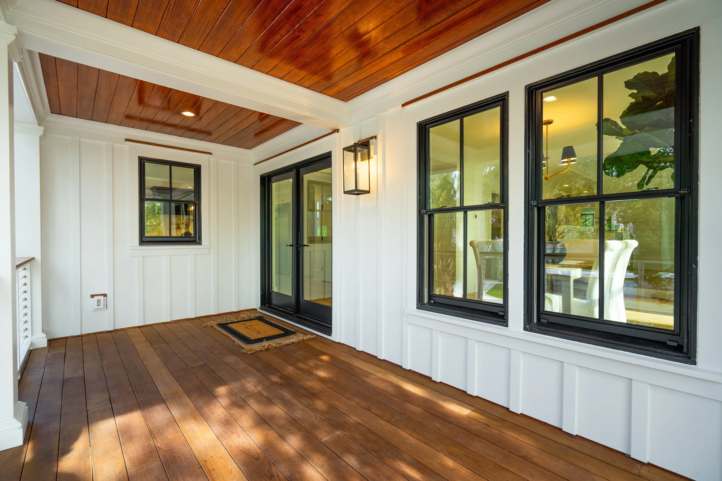 A bright covered porch area with wooden flooring and ceiling, white siding walls, black-framed windows, and a black-framed glass door.