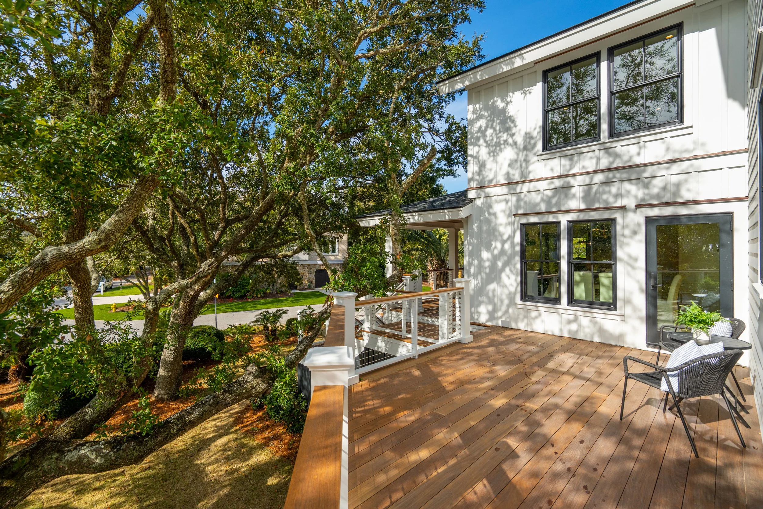 View of a spacious outdoor wooden deck attached to a white house with black-framed windows and glass door, surrounded by lush green trees and a clear blue sky.