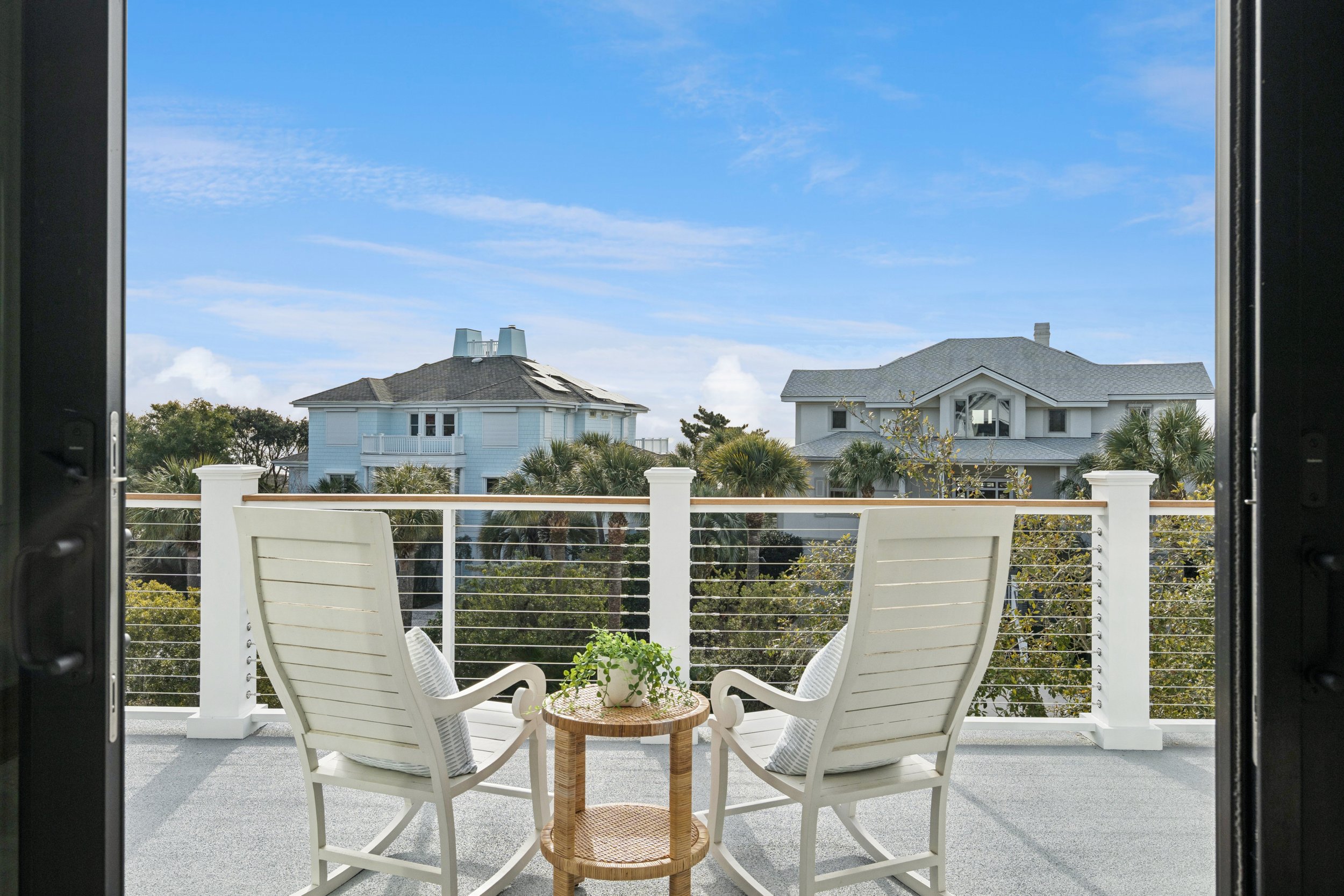 Balcony view of two white chairs and a small table with a potted plant, overlooking neighboring houses and palm trees under a blue sky.