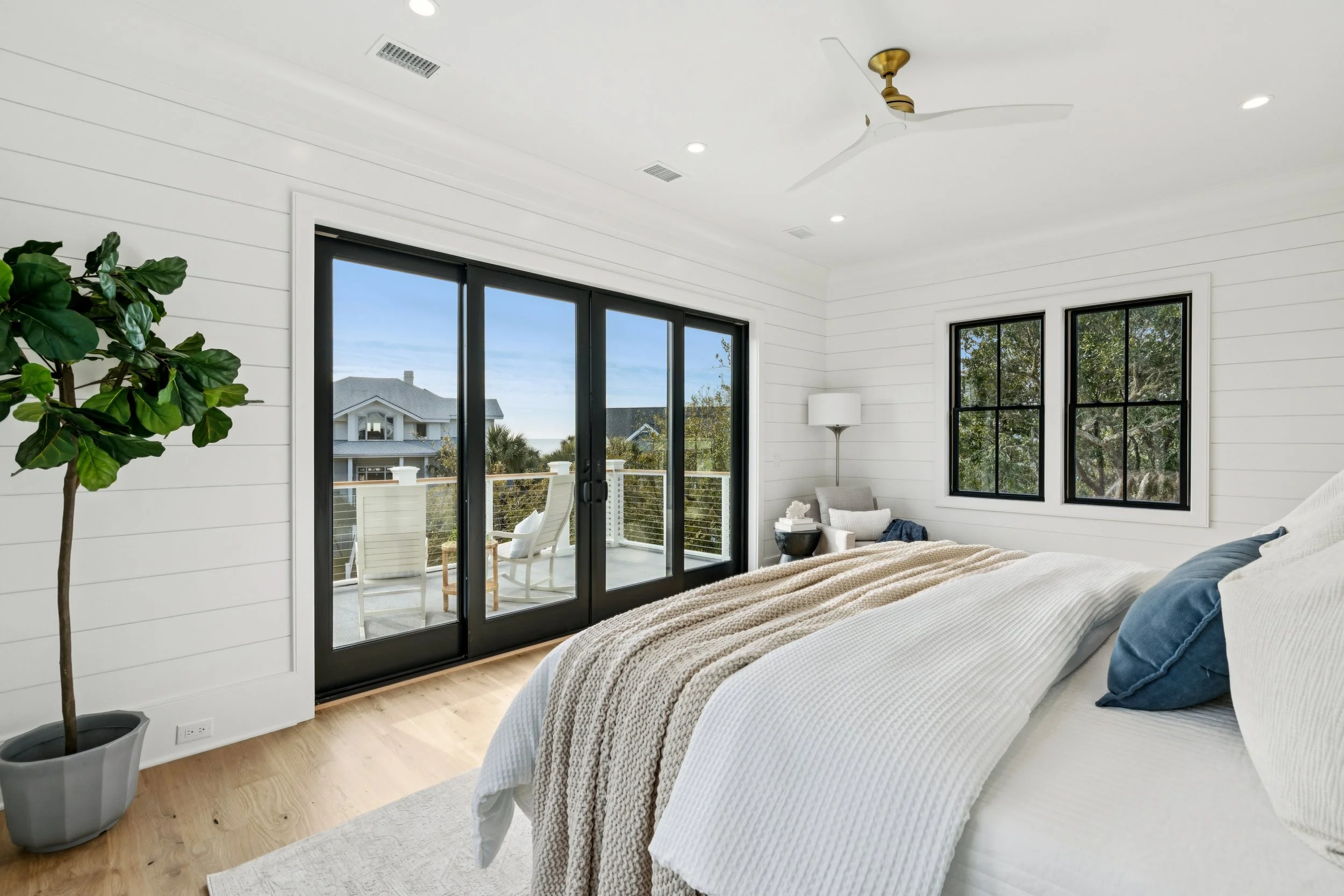 Bright bedroom with white walls, large sliding glass door leading to balcony, and two windows overlooking trees. Includes a bed with white bedding and blue pillows, a potted plant, and a sitting area with a lamp and chair.