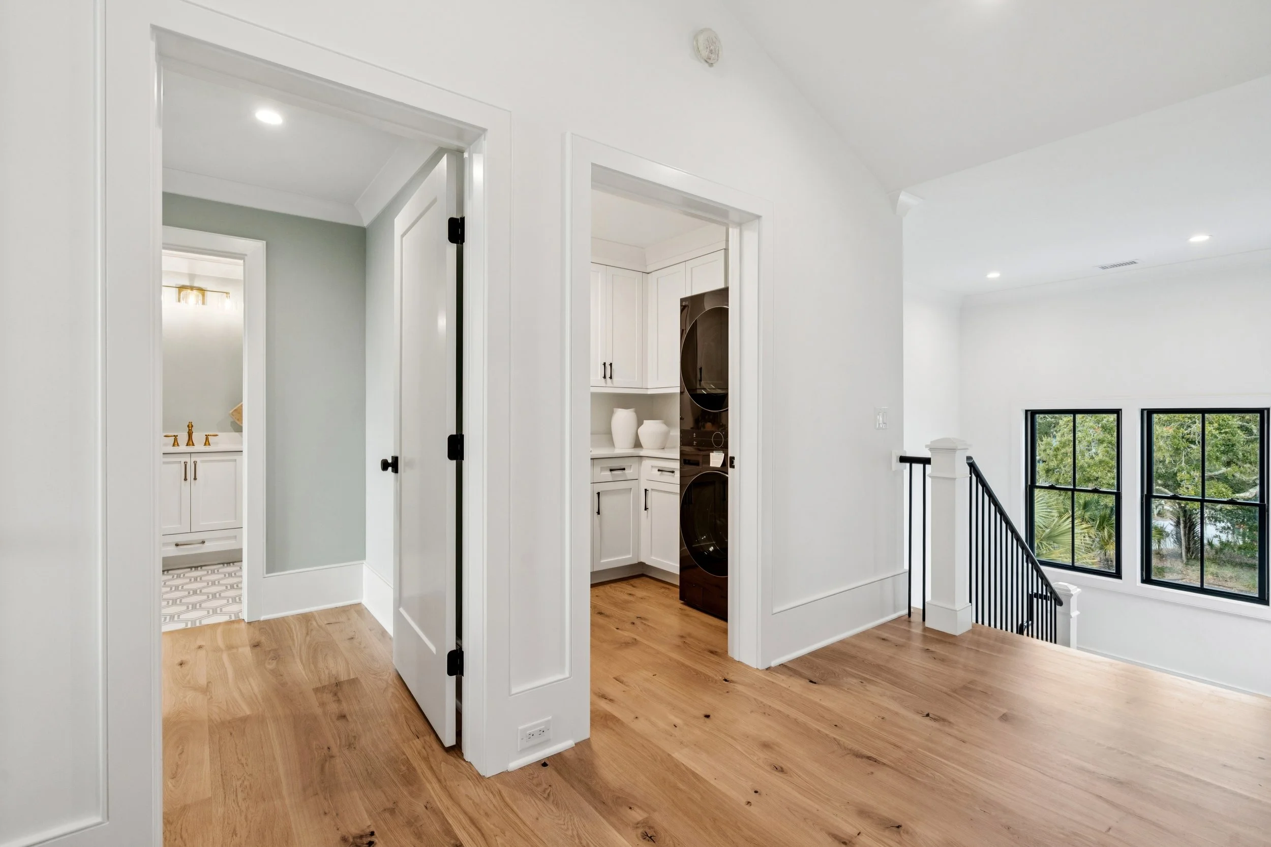 Interior view of a bright, modern home with wooden flooring, white walls, and black window frames, showcasing a staircase and laundry area.