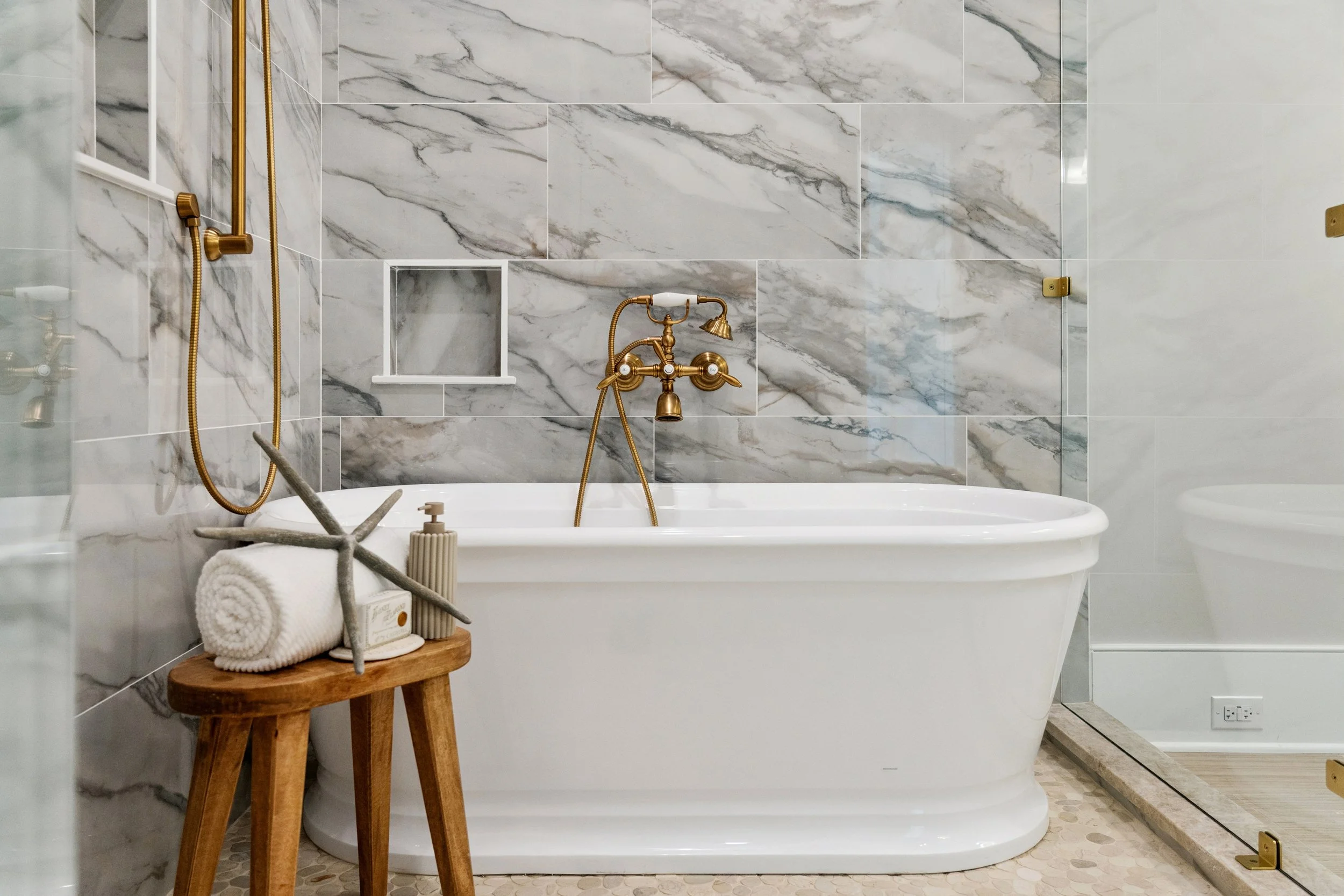 A bathroom with a white bathtub, gold fixtures, marble walls, and a small wooden stool with towels and decor.