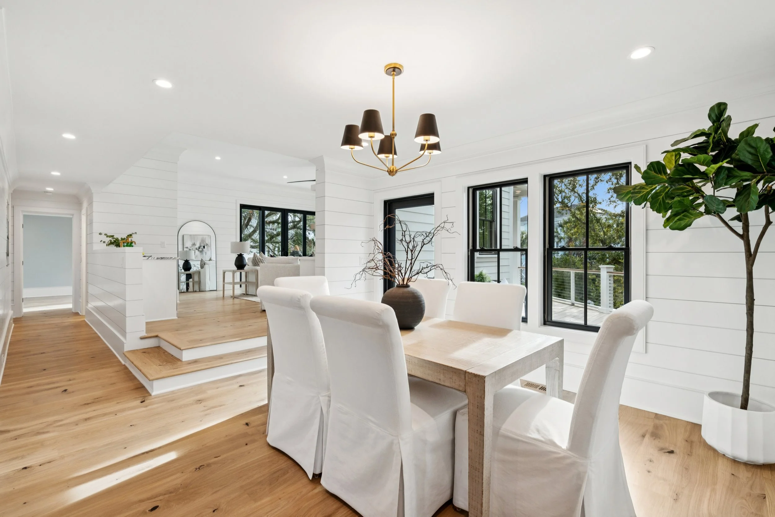 Bright dining room with white walls, pine flooring, a wooden dining table with white chairs, black window frames, a large plant in a white pot, and a black and gold chandelier.
