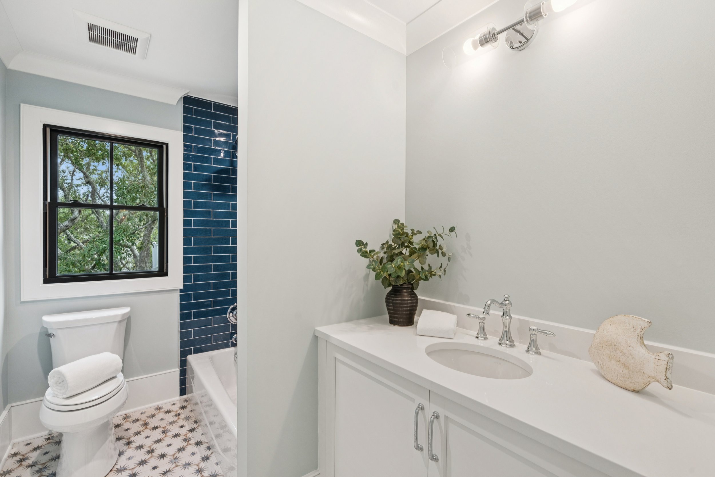 Bathroom with a white vanity, green plant, fish sculpture, and a window showing trees, with a bathtub and blue tiled section in the background.