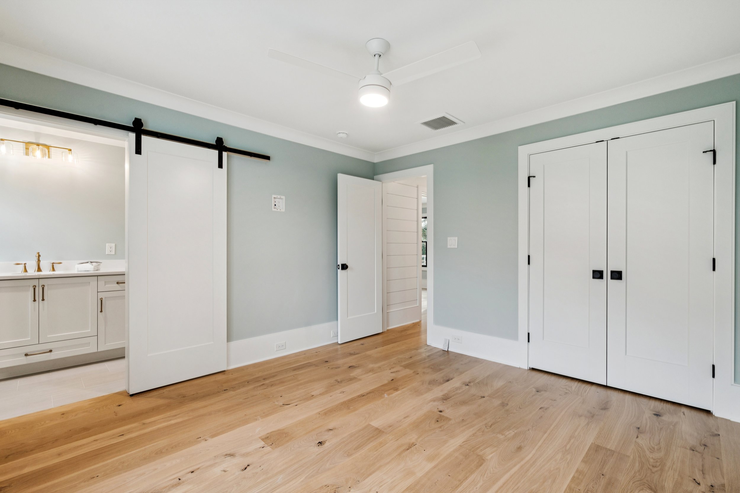Empty room with light green walls, wood floor, white doors, and a visible bathroom with a vanity and gold fixtures.