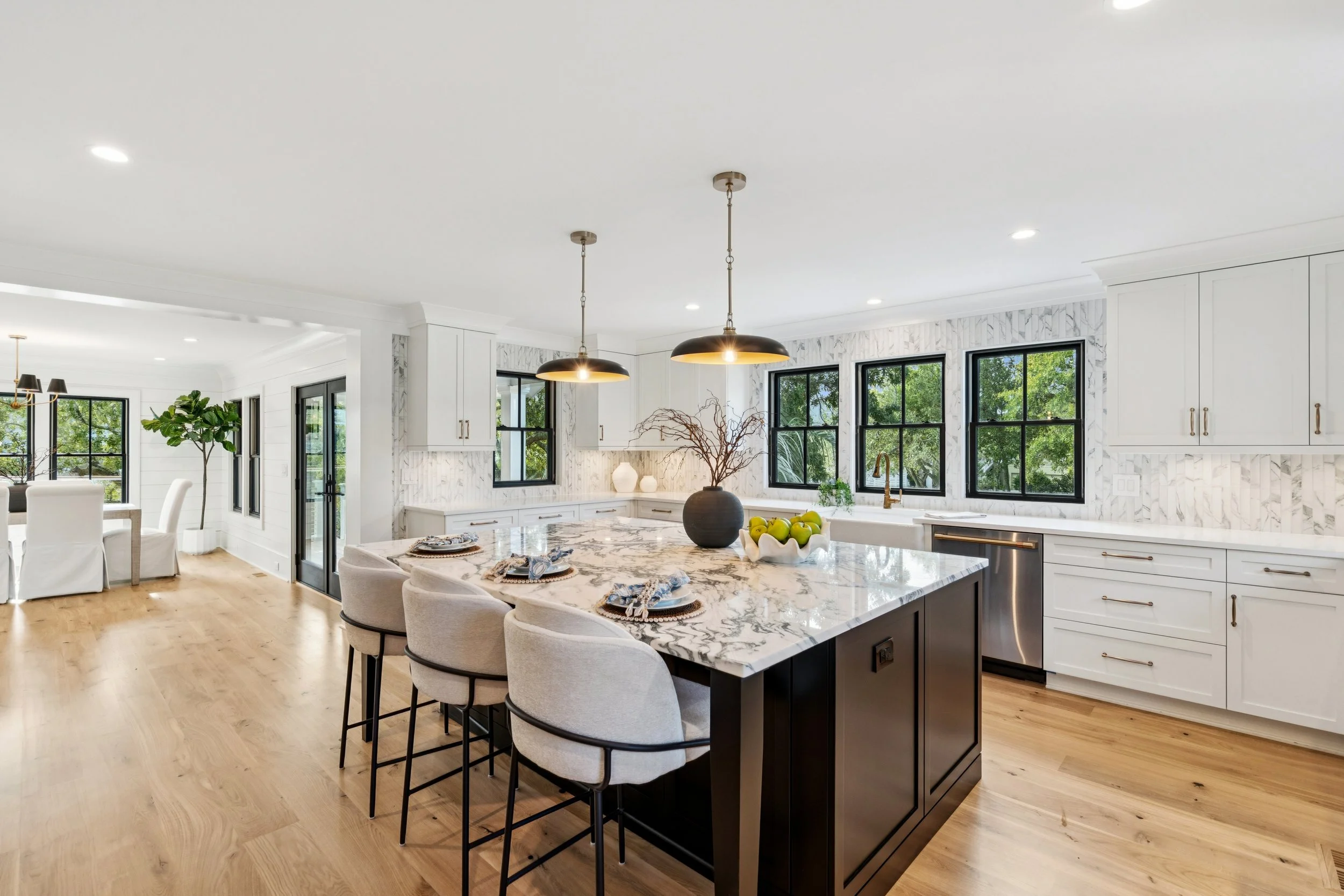 Modern kitchen with white cabinetry, black-framed windows, marble countertops, and a central island with three beige chairs. The island has a decorative vase with branches and a bowl of green apples. Adjacent dining area with white chairs and large w