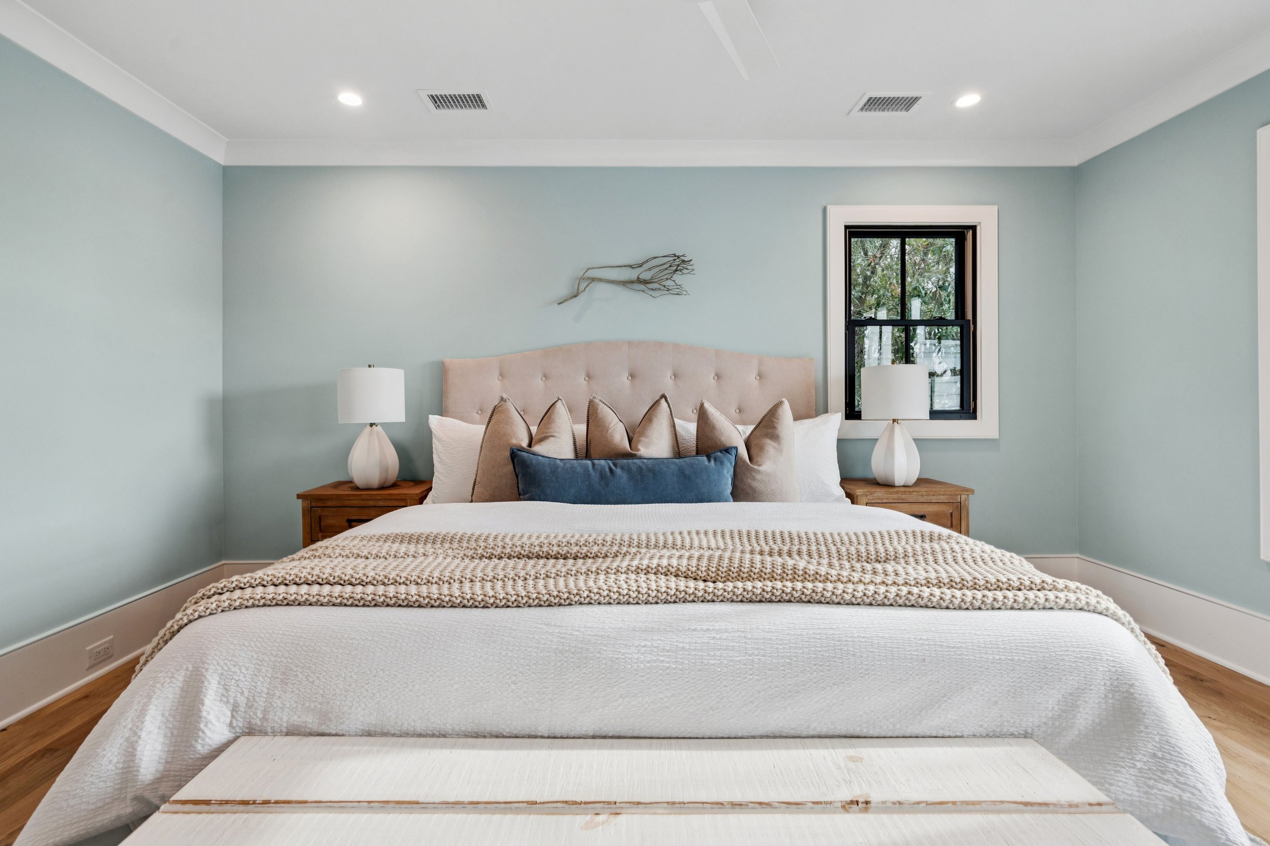 A neatly made bed with a padded beige headboard, decorative pillows including beige, white, and a central blue pillow, two matching lamps on wooden nightstands, a window with black trim, light blue walls, and a piece of driftwood art above the bed.