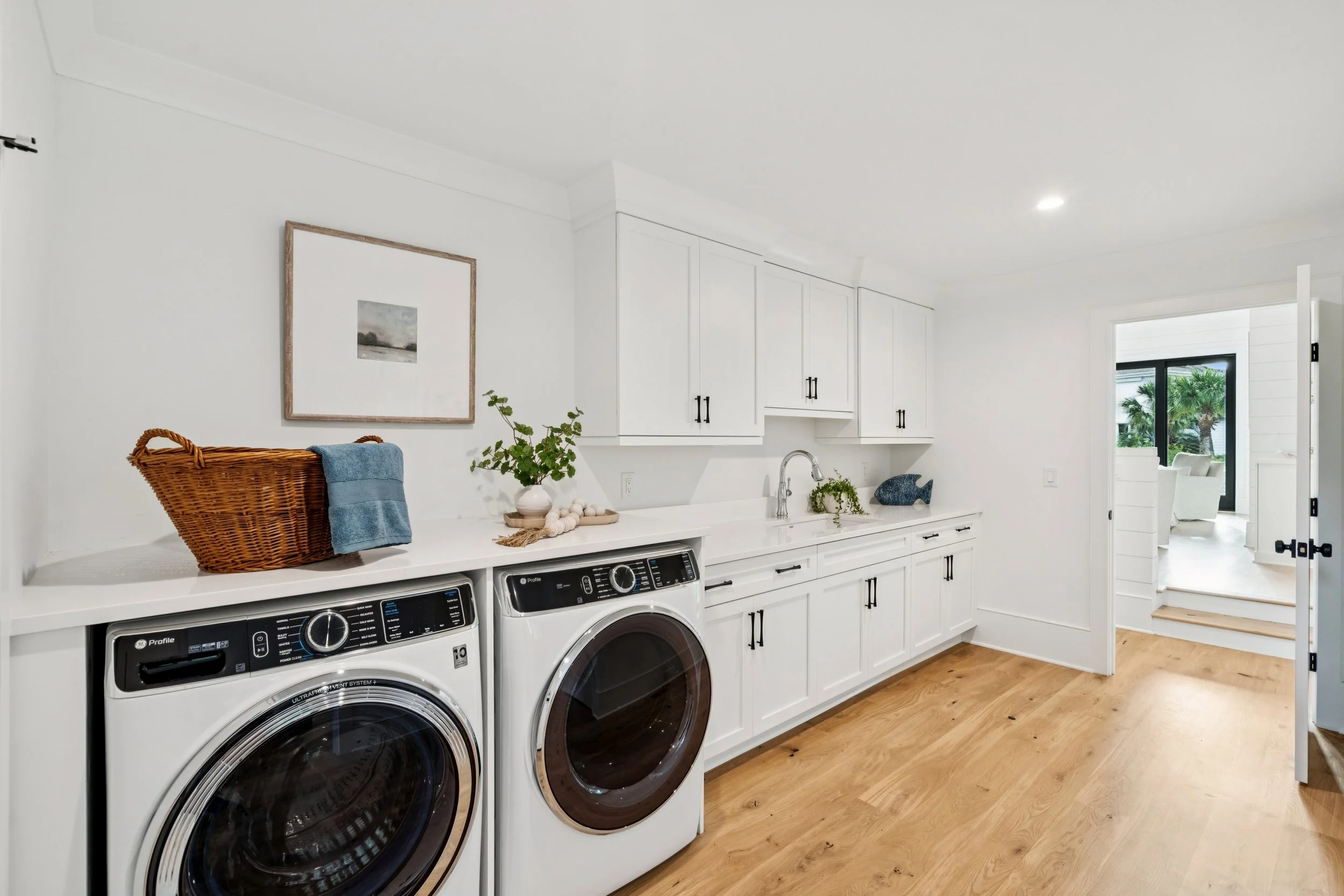 Laundry room with white cabinets, a countertop with a basket, plant, seashells, decorative fish, and a washer and dryer, with a view into a bright sitting area outside.