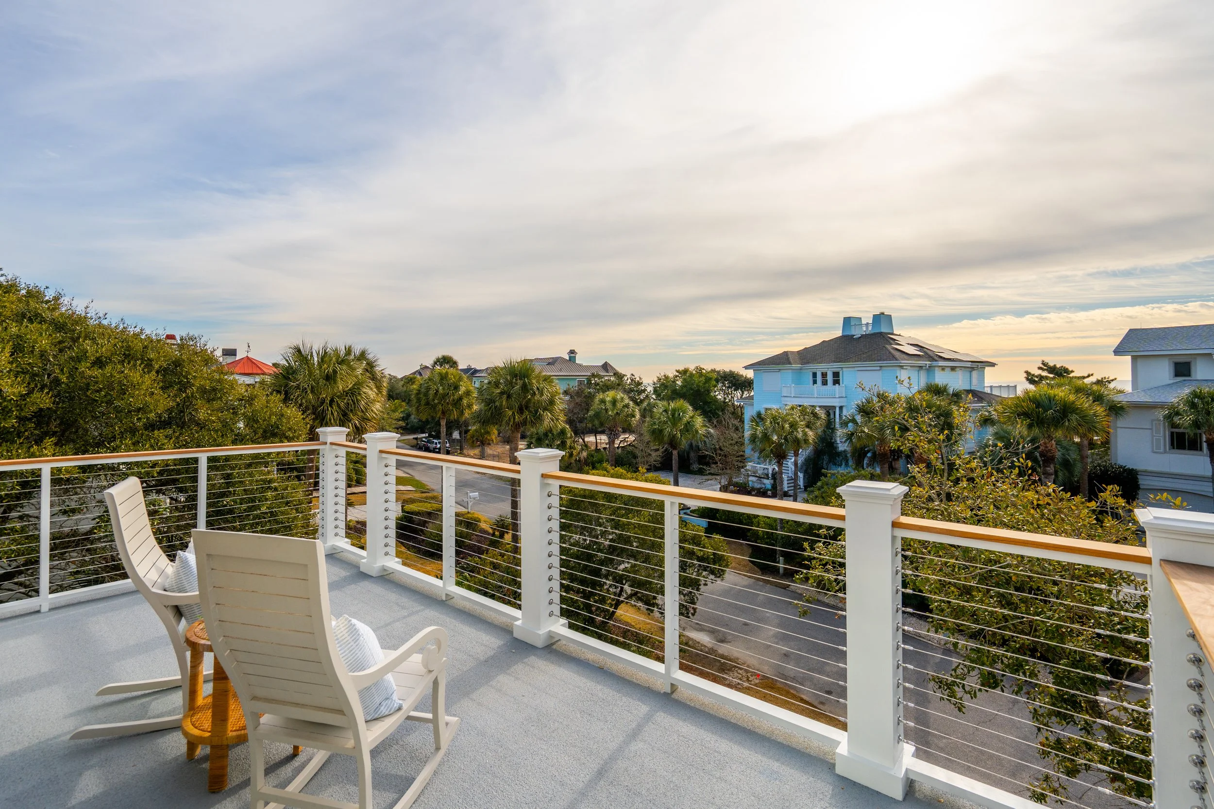 View from a balcony with white rocking chairs overlooking palm trees, houses, and a partly cloudy sky