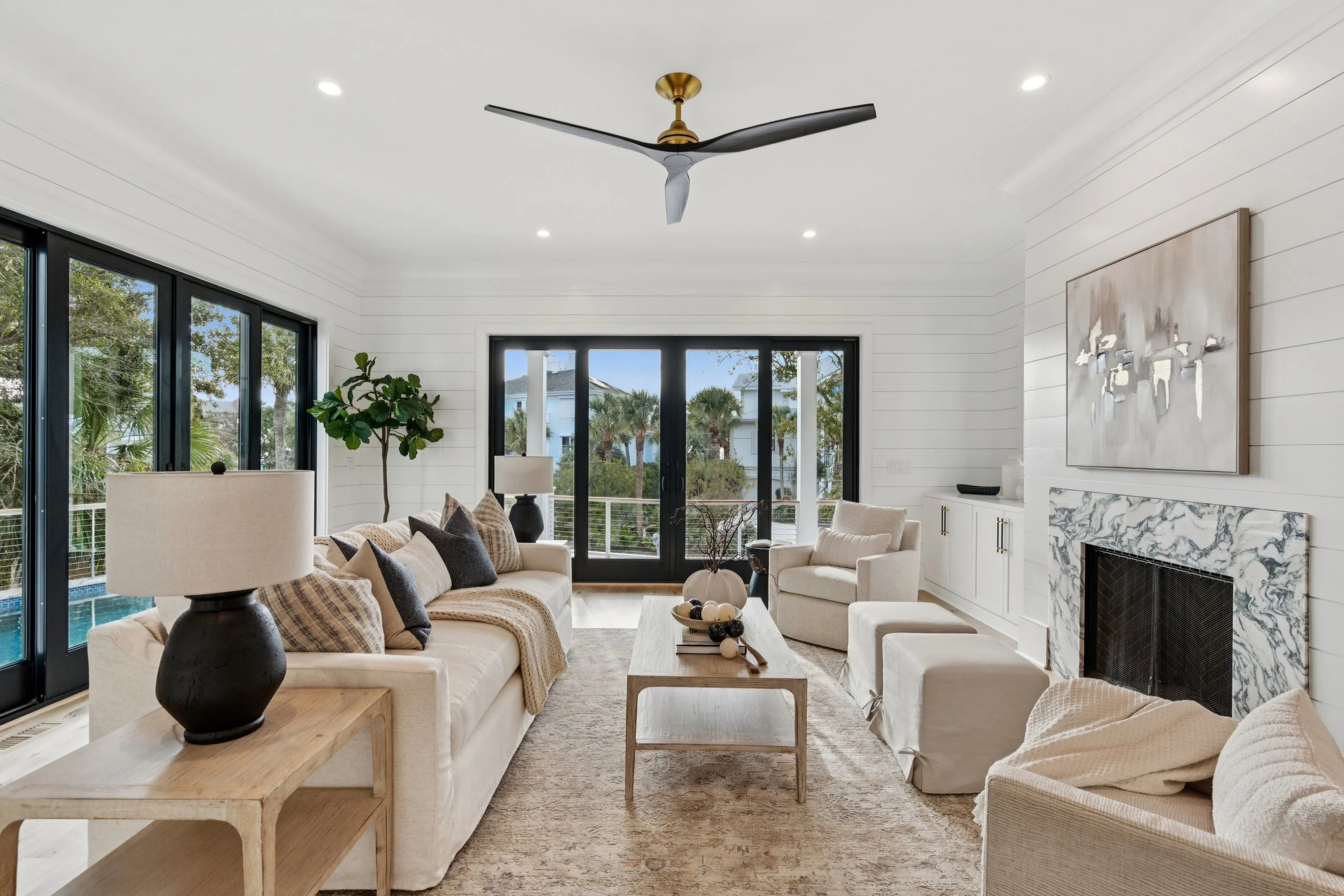 Bright living room with cream sofas, a wooden coffee table, and large glass doors leading to a balcony with trees outside.