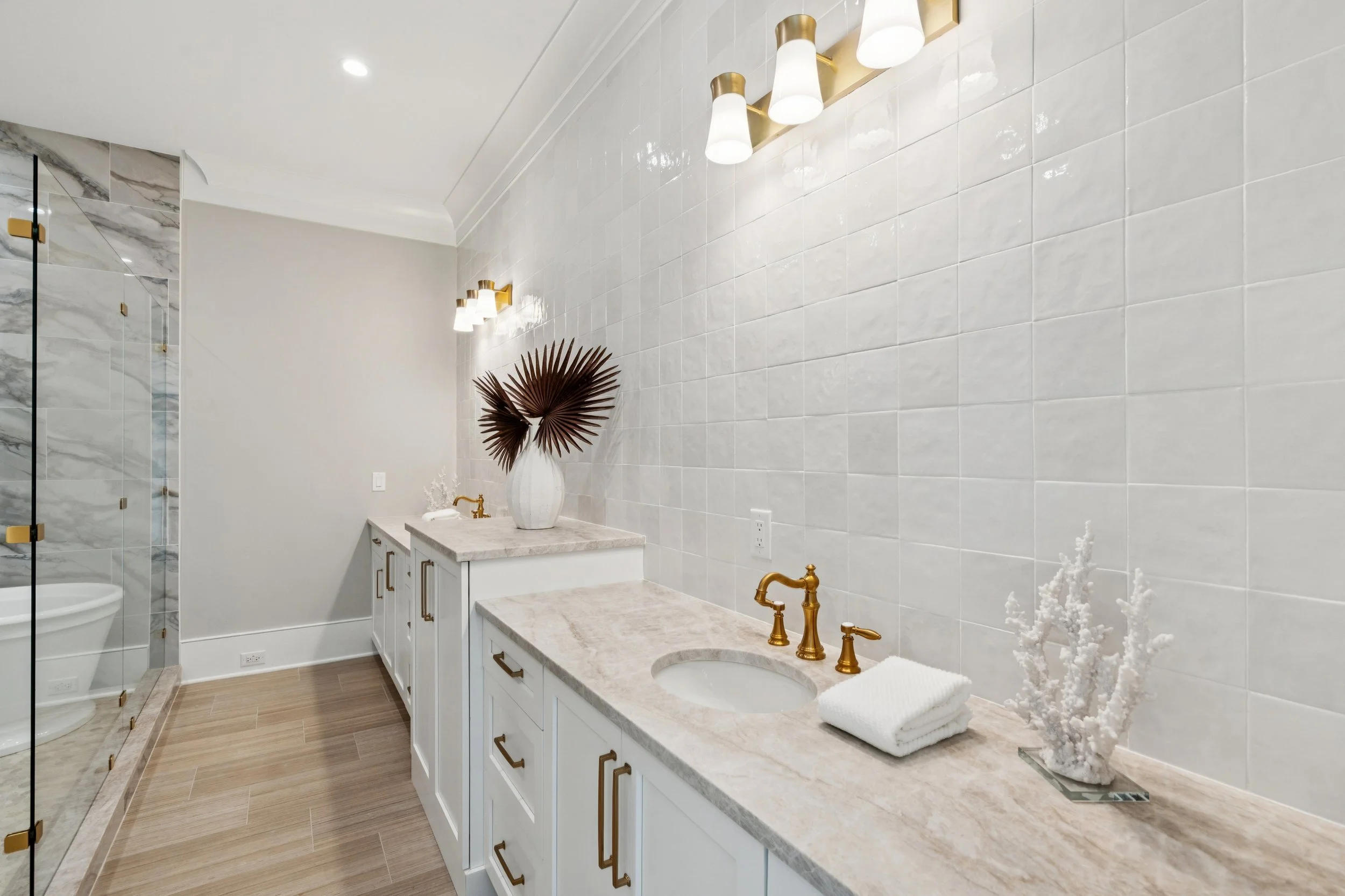 Modern bathroom with beige marble countertops, white cabinets with gold handles, gold fixtures, decorative vase with dried leaves, folded white towels, white tiled wall, and a glass shower with marble walls.