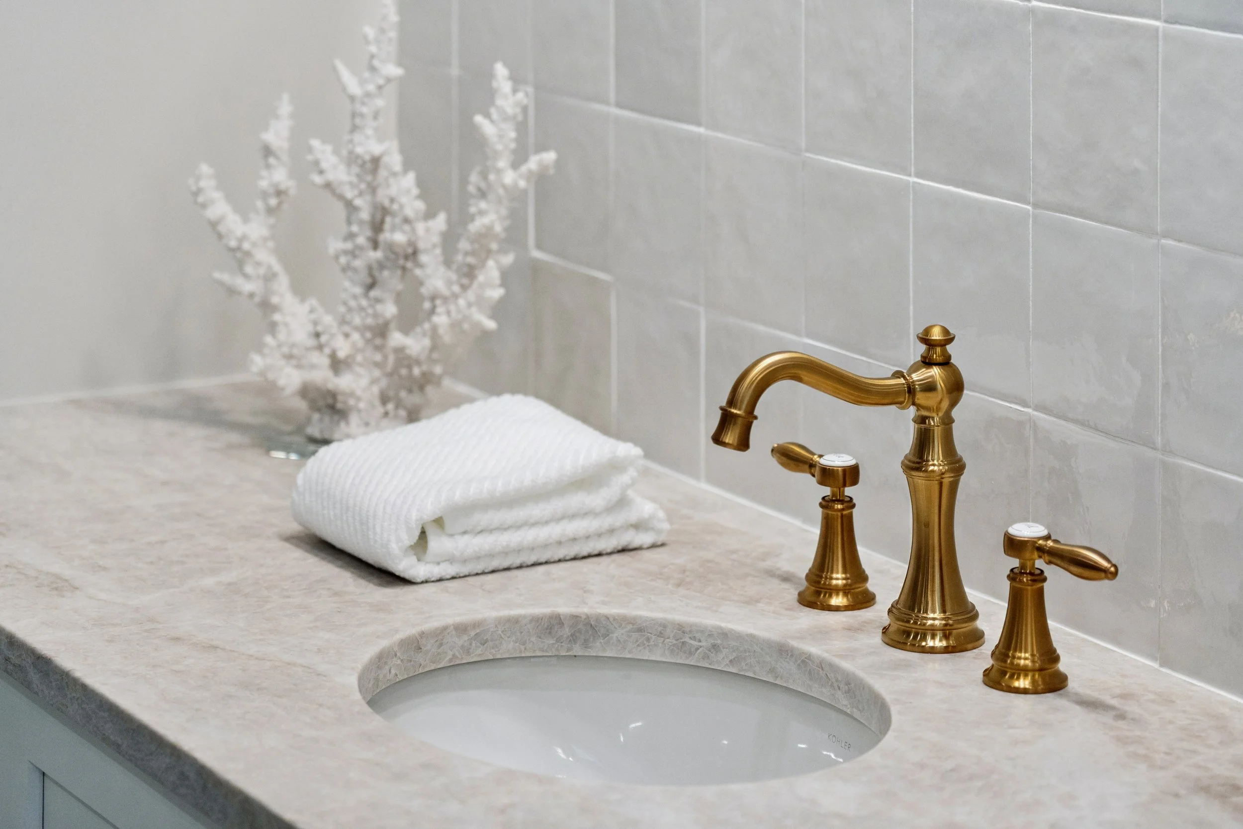 Bathroom sink with brass faucet, white folded towel, decorative white coral. The sink is in a beige marble countertop with a light gray tiled wall behind it.