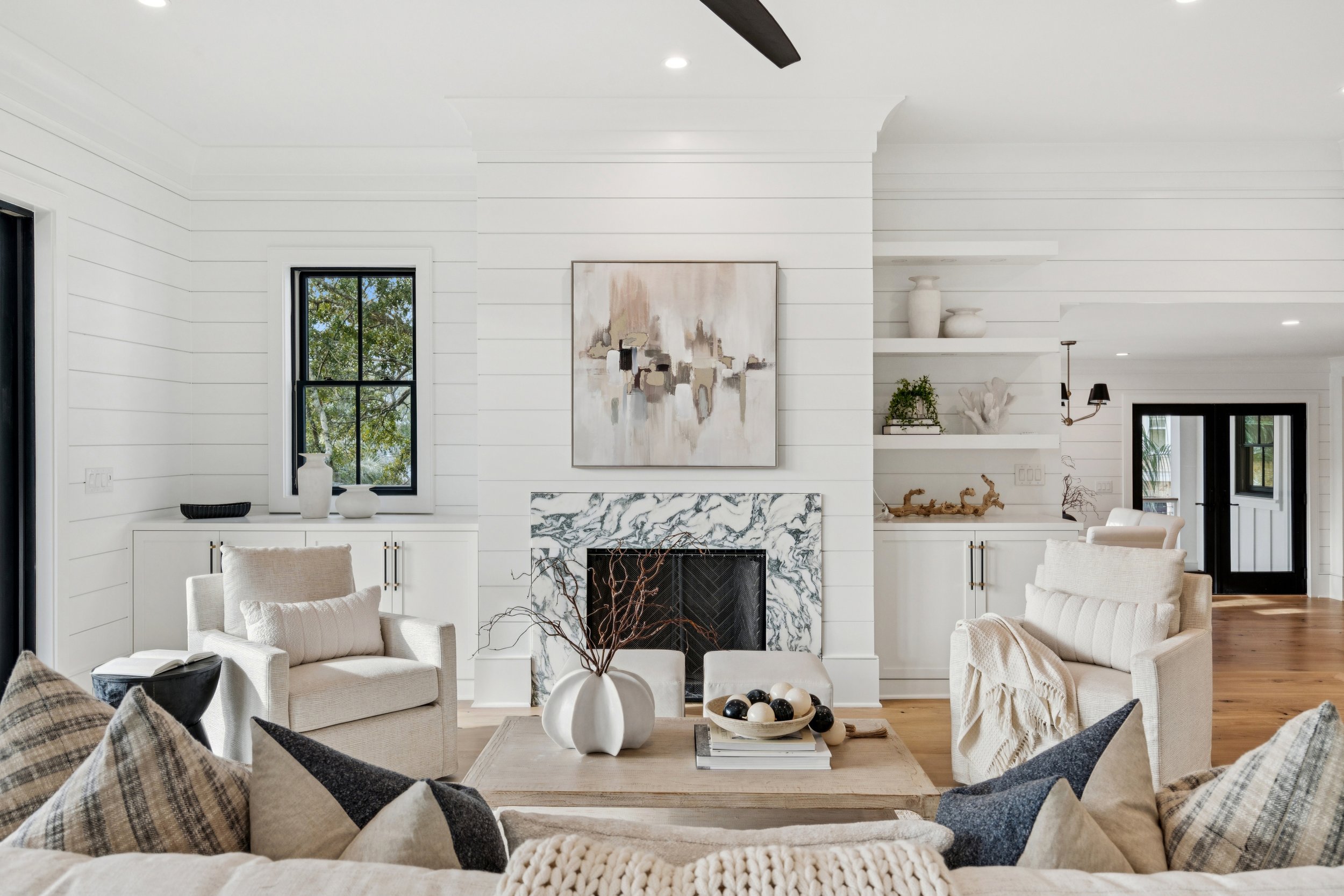 Living room with white walls, a marble fireplace, neutral sofas, decorative pillows, a coffee table with a vase and decorative balls, and black framed windows.