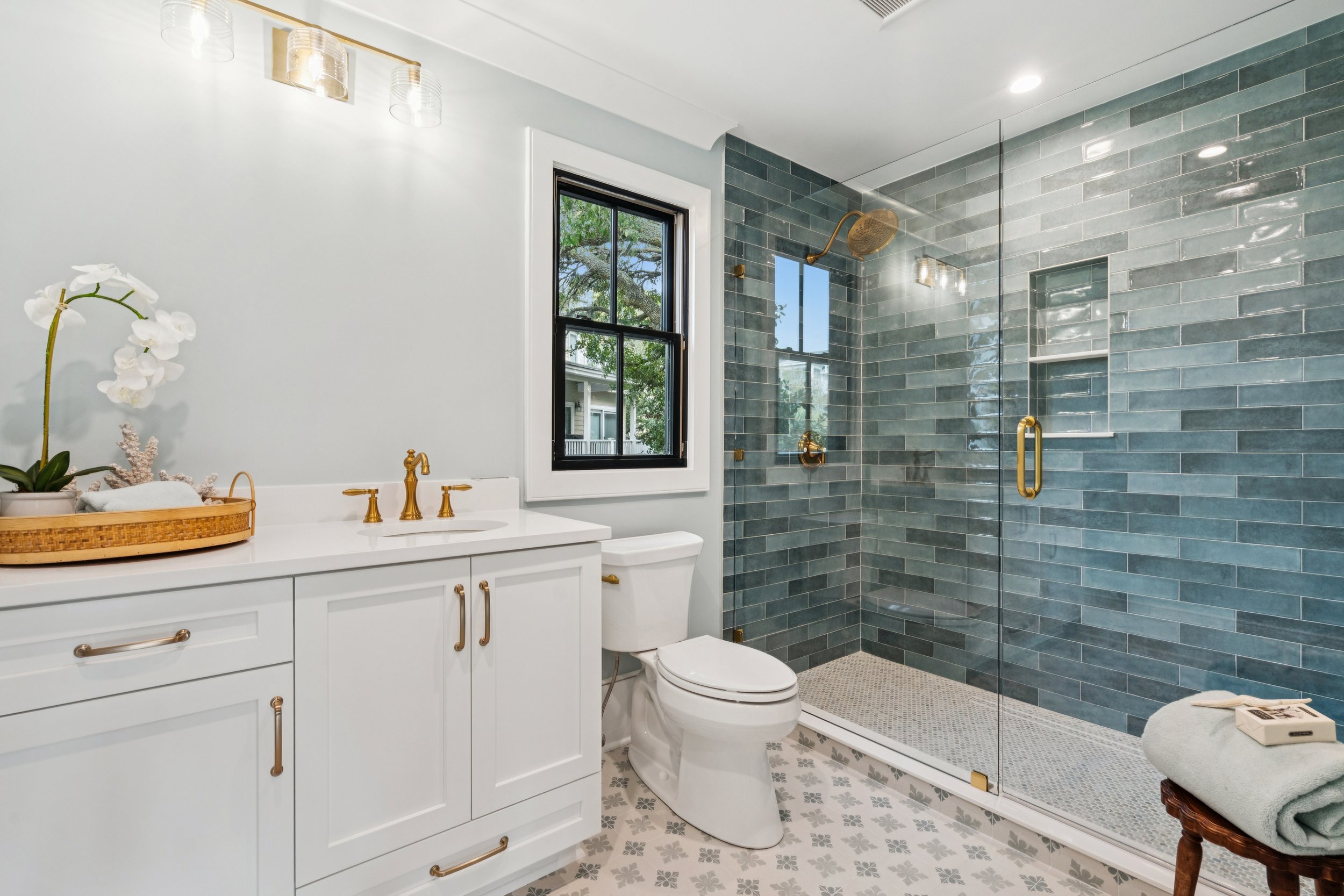 Modern bathroom with white vanity, gold fixtures, black framed window, and blue tile shower with glass door, decorative floor tiles, and a window view of trees outside.