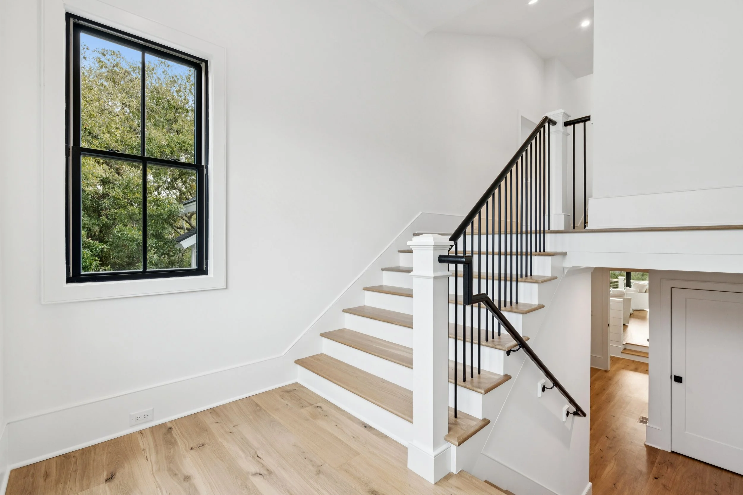 Bright, modern interior staircase with light wood steps, white risers, black metal railing, and large window showing green trees outside.