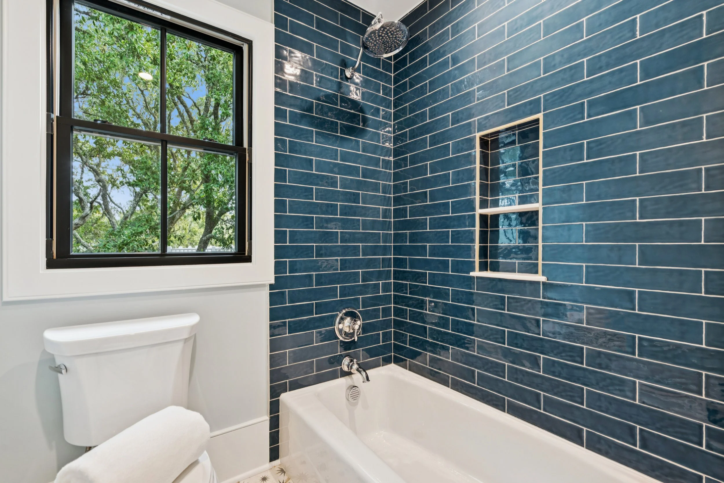 Bathroom with blue subway tile walls, a white bathtub, a black-framed window showing green trees outside, a shower head, and a small built-in shower shelf.