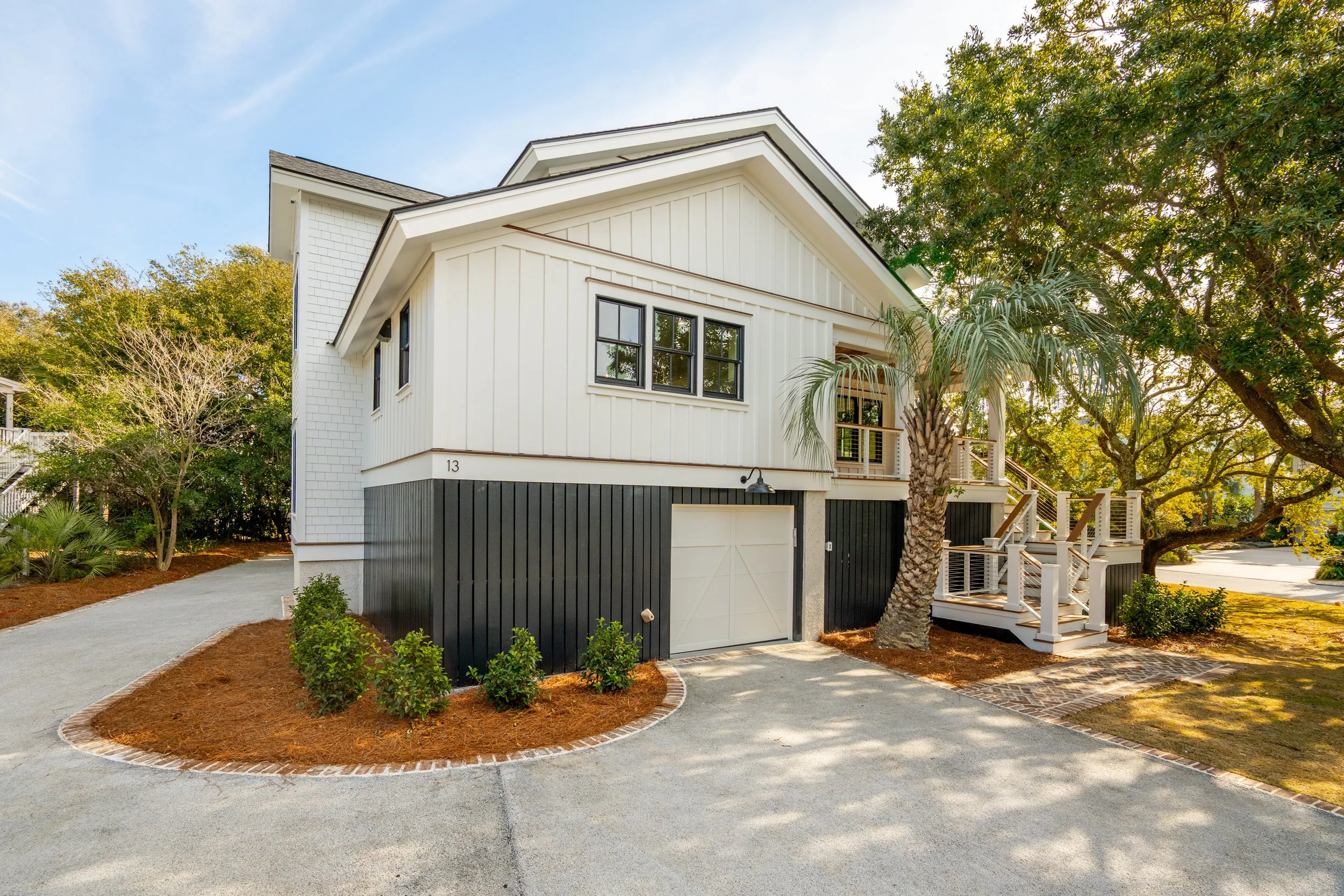 A modern two-story house with white and black exterior, a garage door, wooden stairs, a balcony, and surrounded by trees and landscaping.