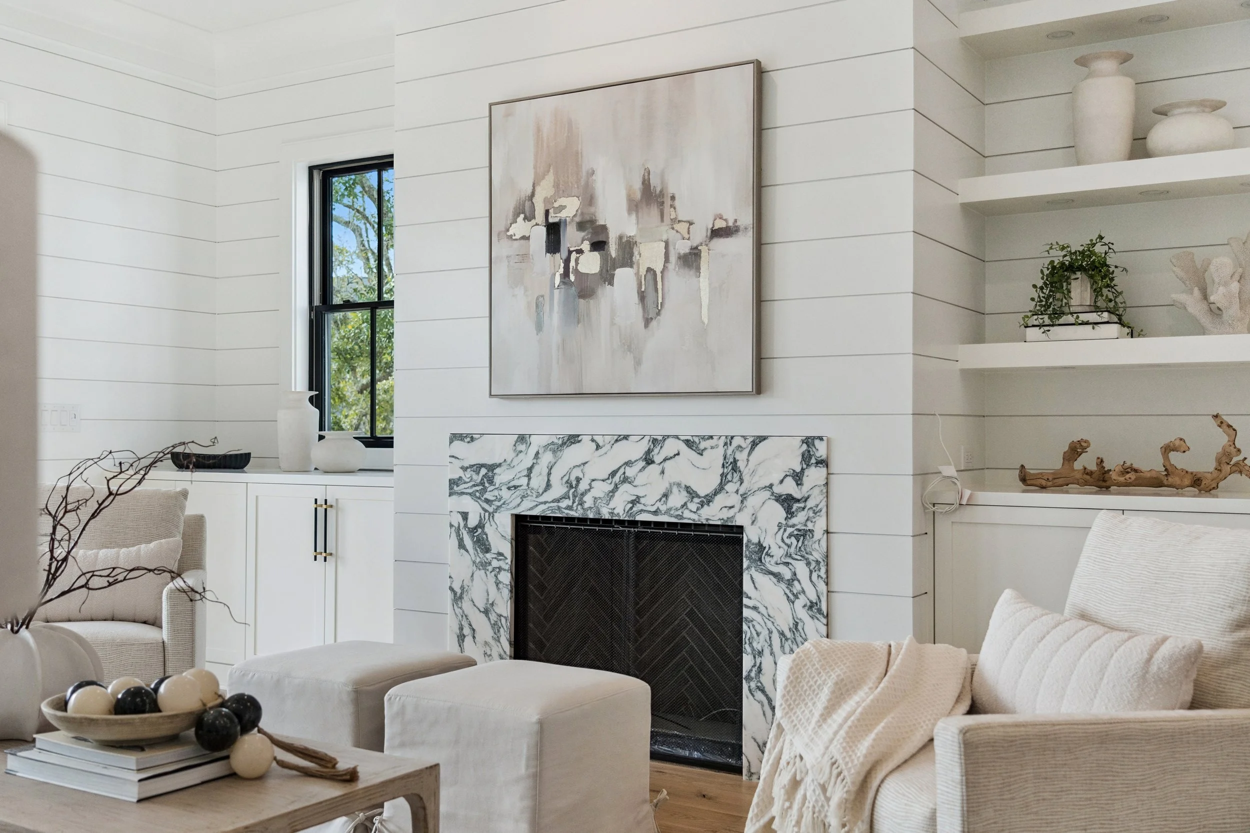Living room with white walls, a abstract painting, a marble fireplace, a window with trees outside, beige furniture, and decorative vases and driftwood.