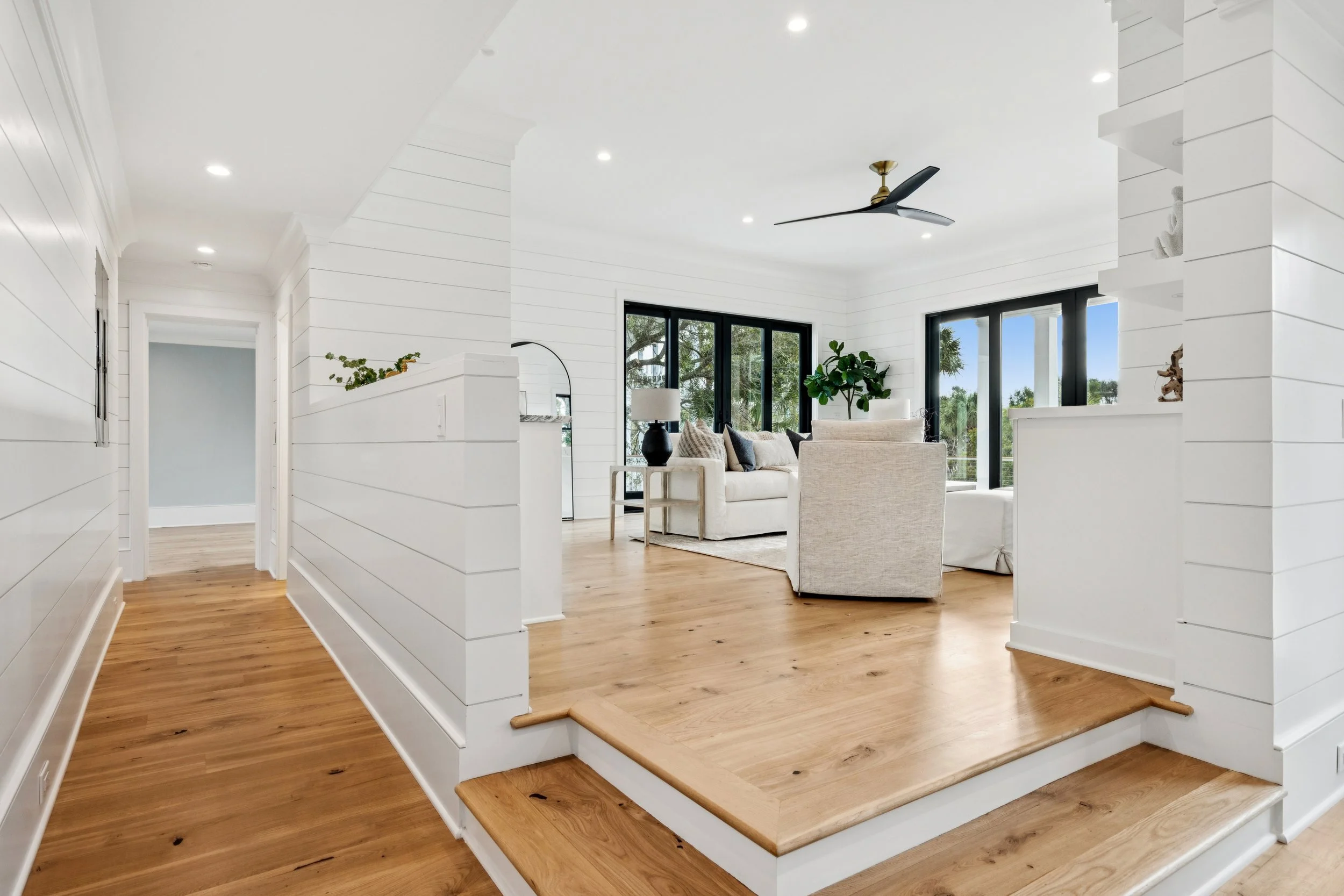 Bright living room with white shiplap walls, hardwood floors, a ceiling fan, black-framed windows, and minimalist furniture including a white sofa and beige armchair.