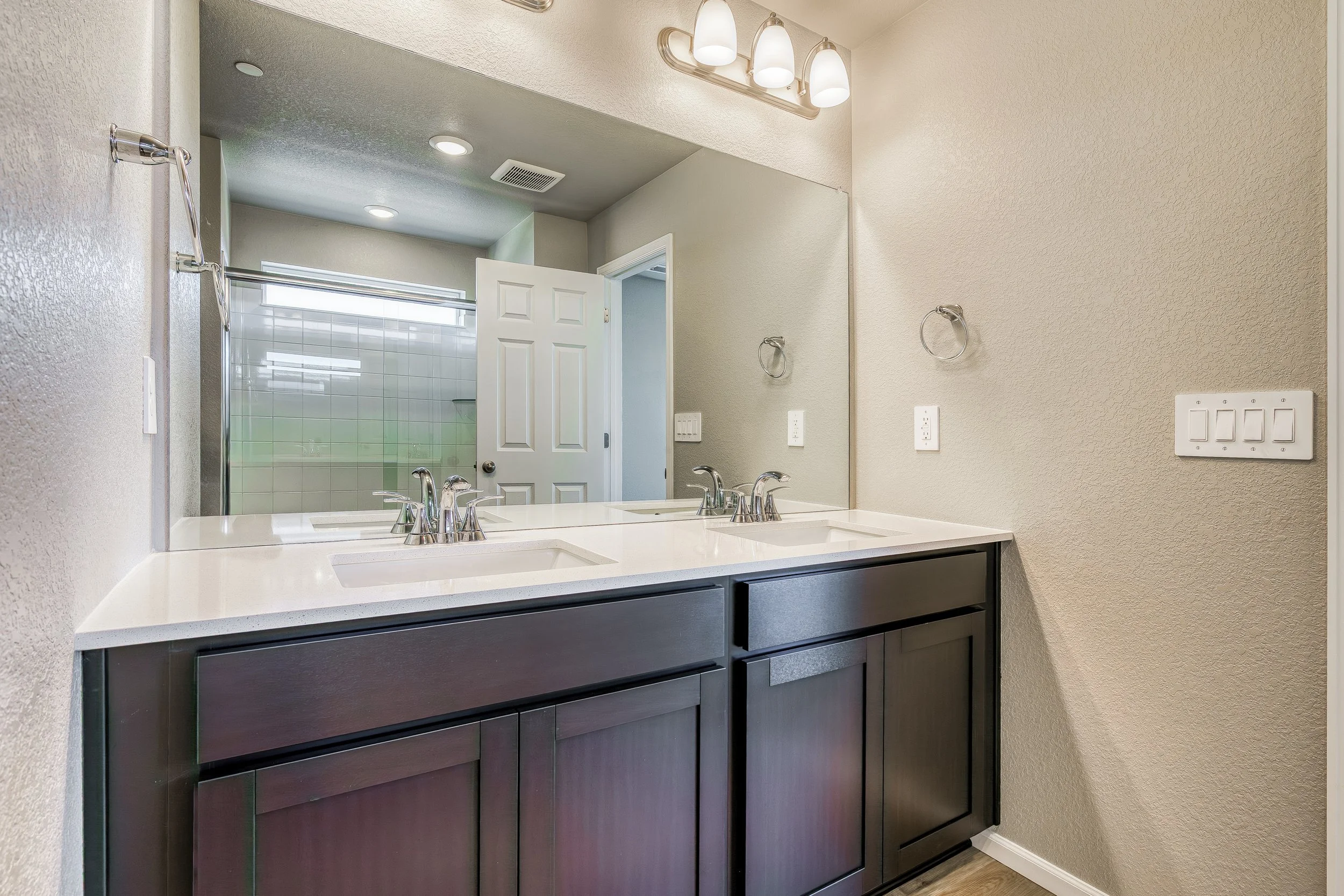 Bathroom with dual sinks, dark wood vanity, large mirror, beige walls, towel rings, and a visible shower with glass door in the background.