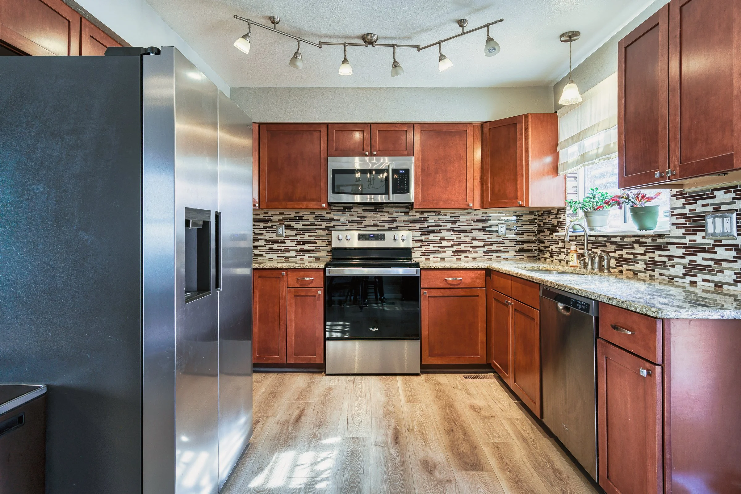 Kitchen with honey-colored wooden cabinets, stainless steel appliances including a refrigerator, microwave, stove, and dishwasher, a mosaic tile backsplash, granite countertops, a window with a shade, and potted plants on the windowsill.