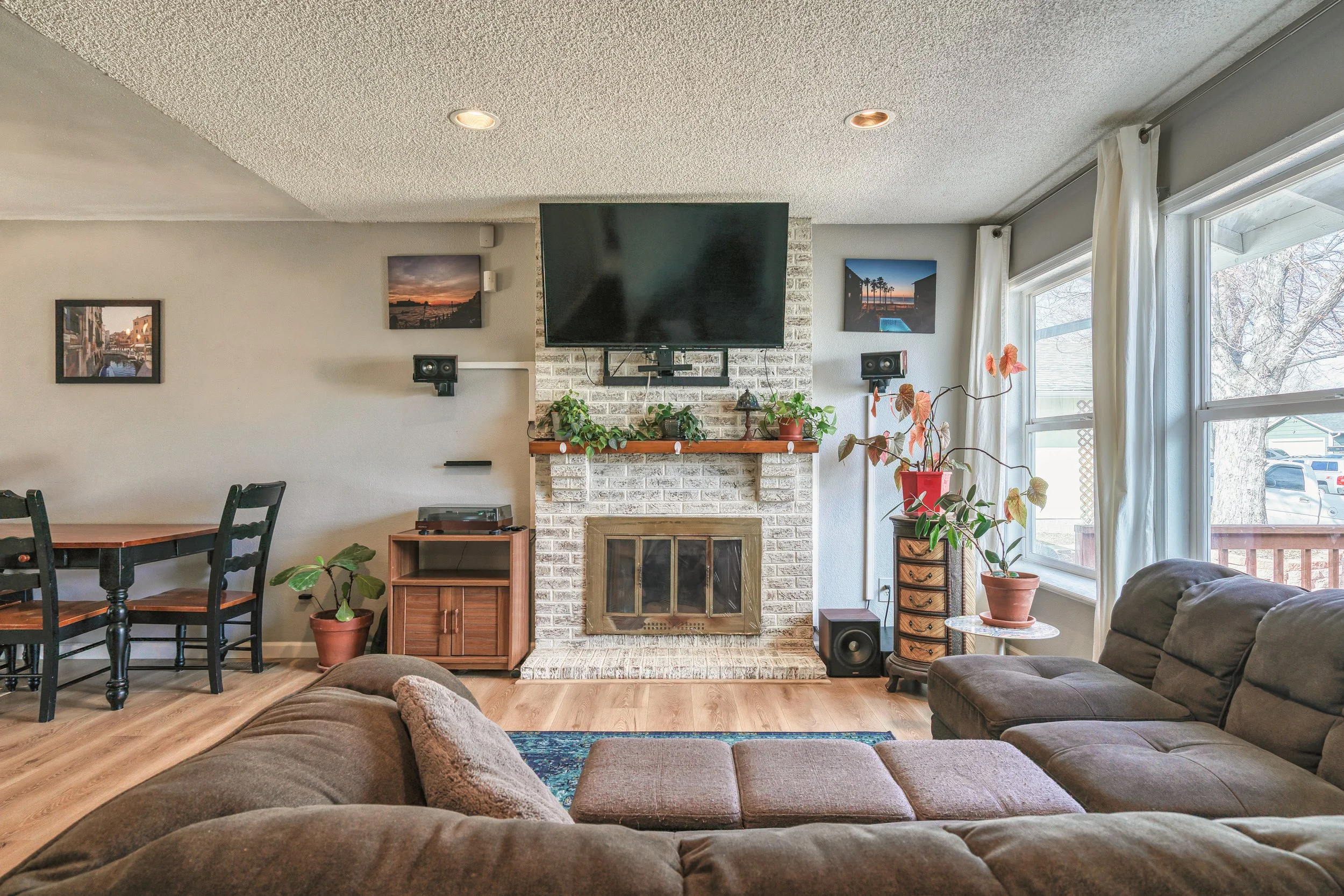 Living room with a brown sectional sofa, a brick fireplace with a TV mounted above, potted plants, windows with white curtains, and wall art.