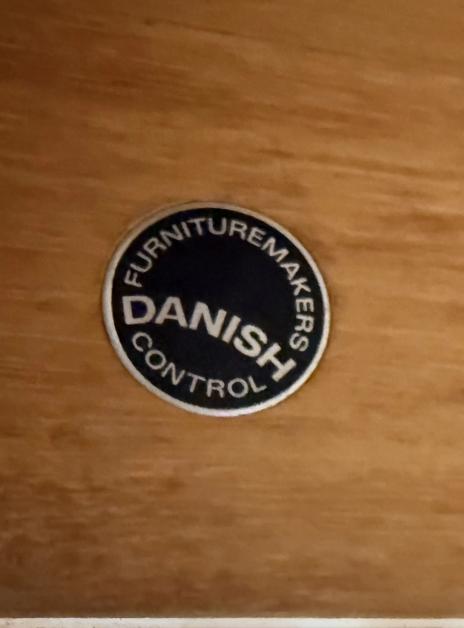 Close-up of a circular black and white sticker on a wooden surface with partially visible text reading 'FURNITUREMAKER DAVIDSON CONTROLE'.