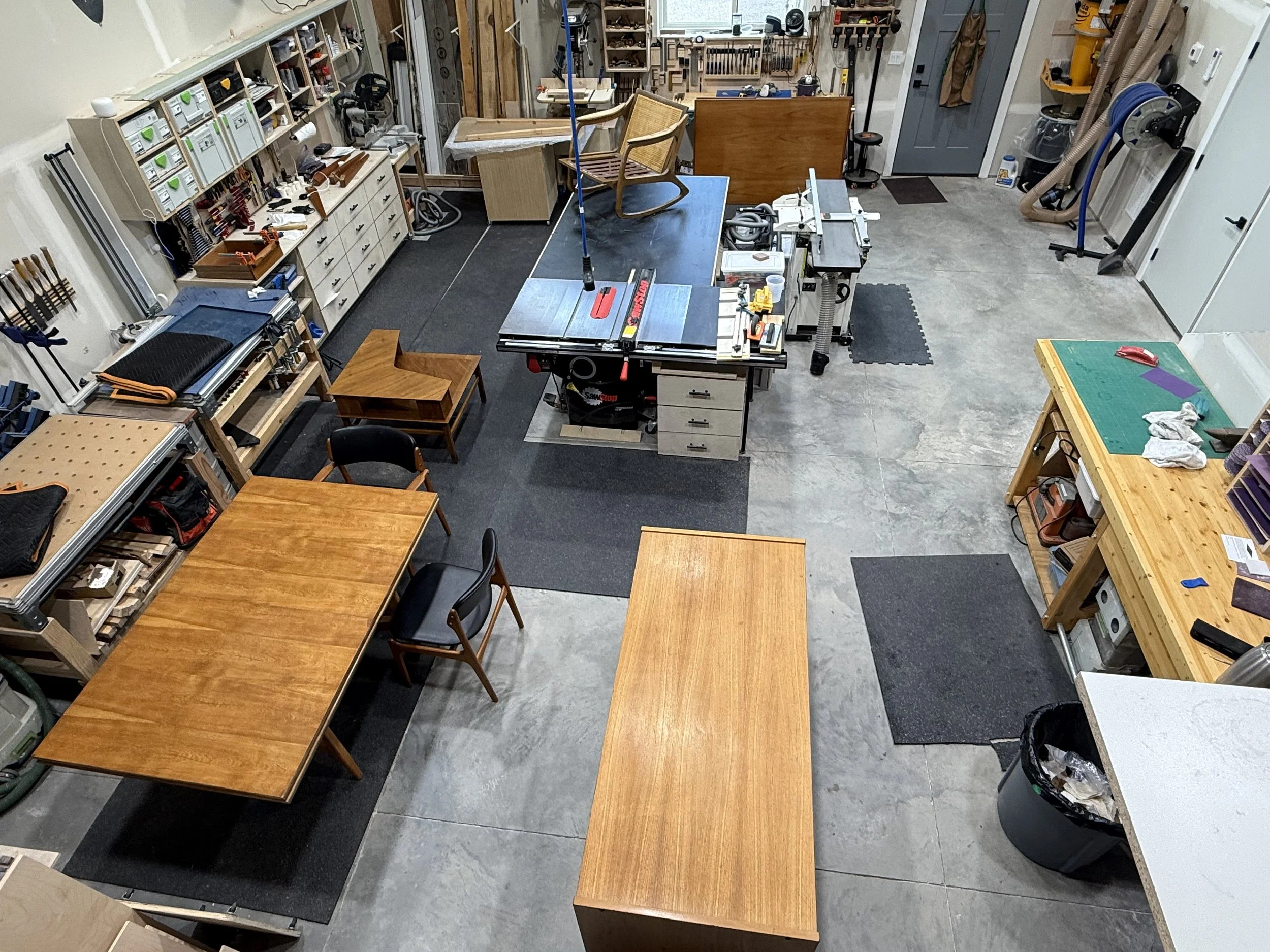 A well-organized woodworking workshop with various tools, work tables, and storage units for woodworking supplies, as well as teak, walnut and rosewood furniture undergoing, restoration, repair or refinishing.