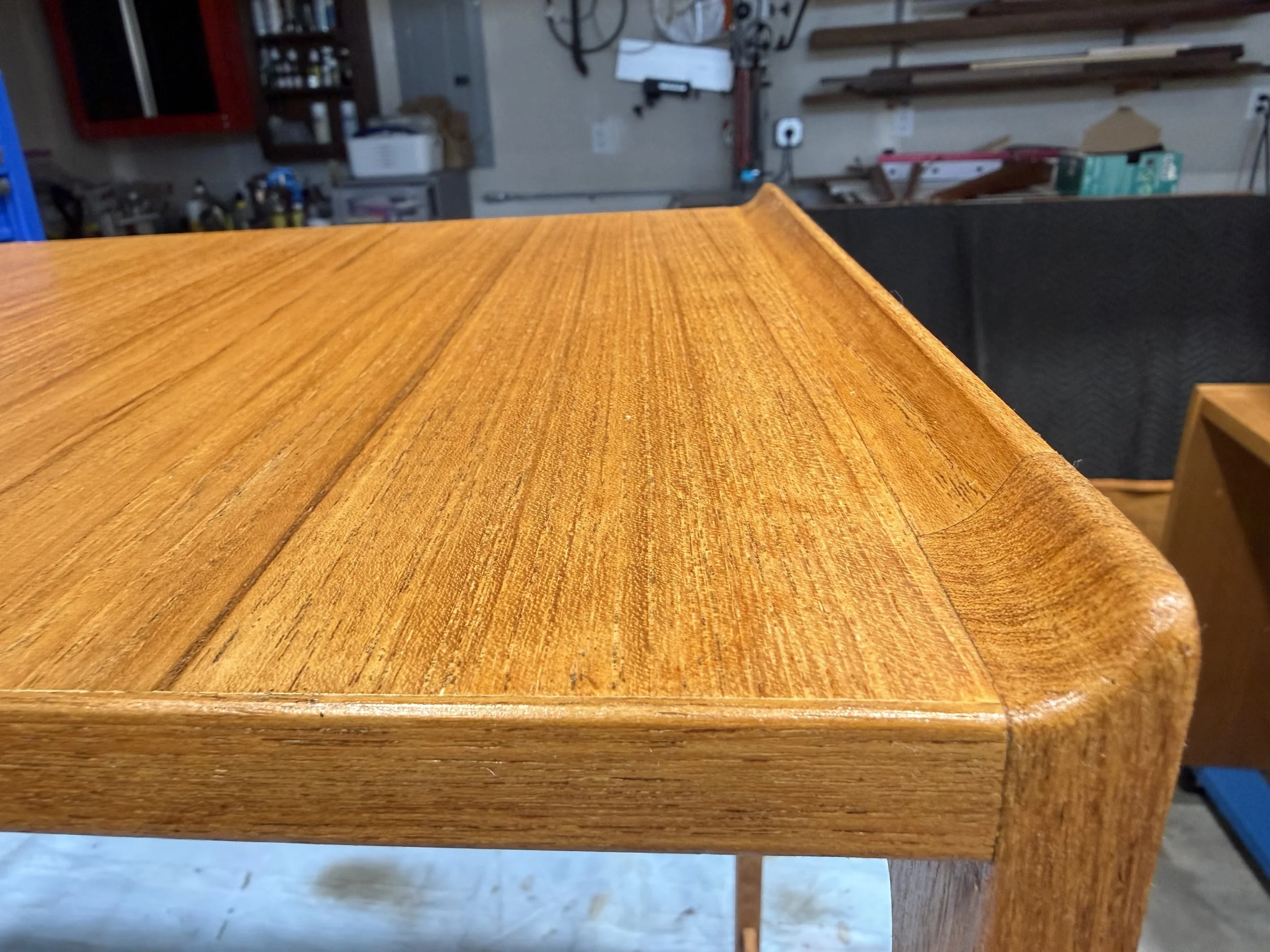 Close-up of a wooden table with a smooth finish, showing the grain and corner detail, in a workshop or garage setting.