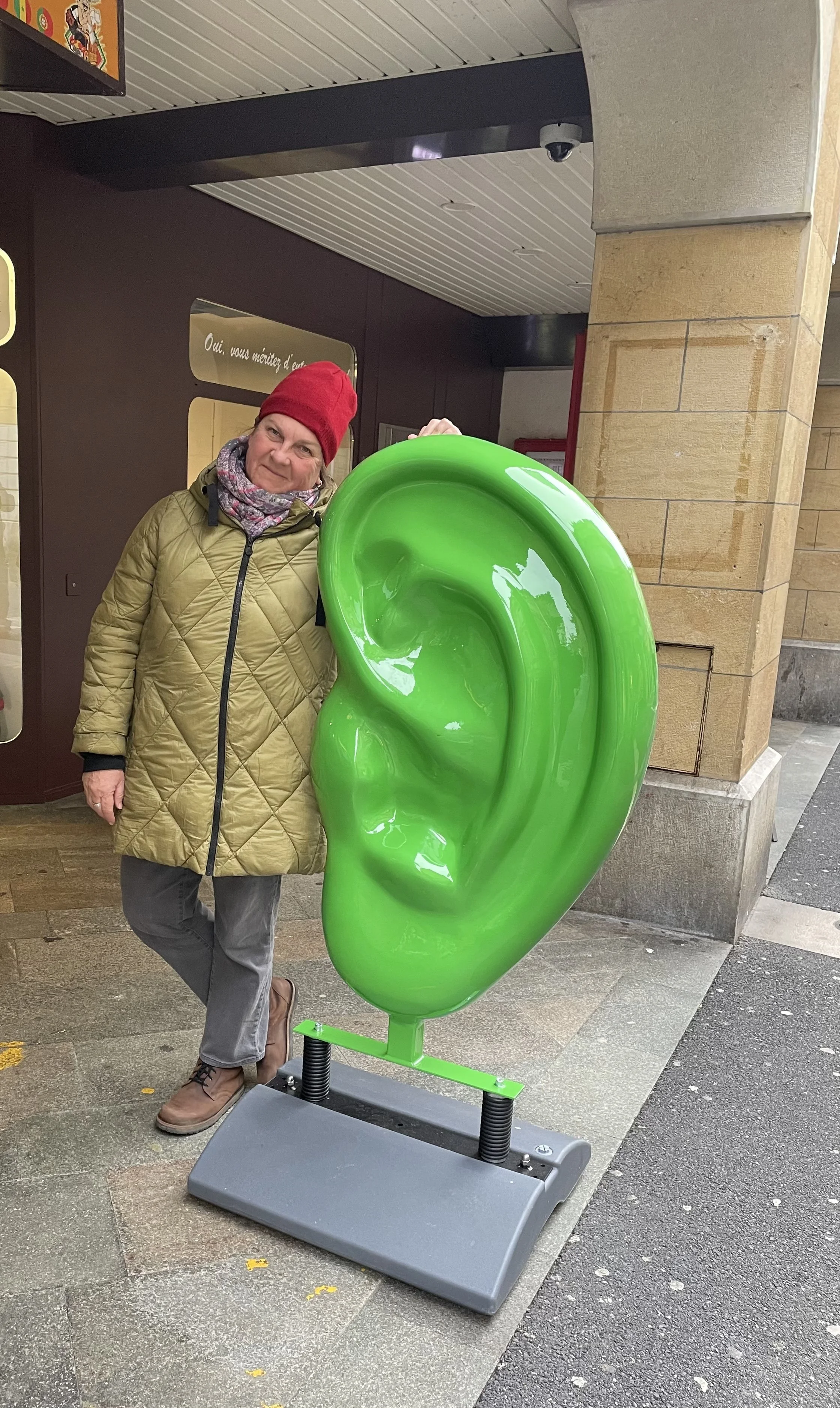 A woman in a tan quilted coat, red beanie, and scarf standing next to a large, green sculpture of an ear outside a building.