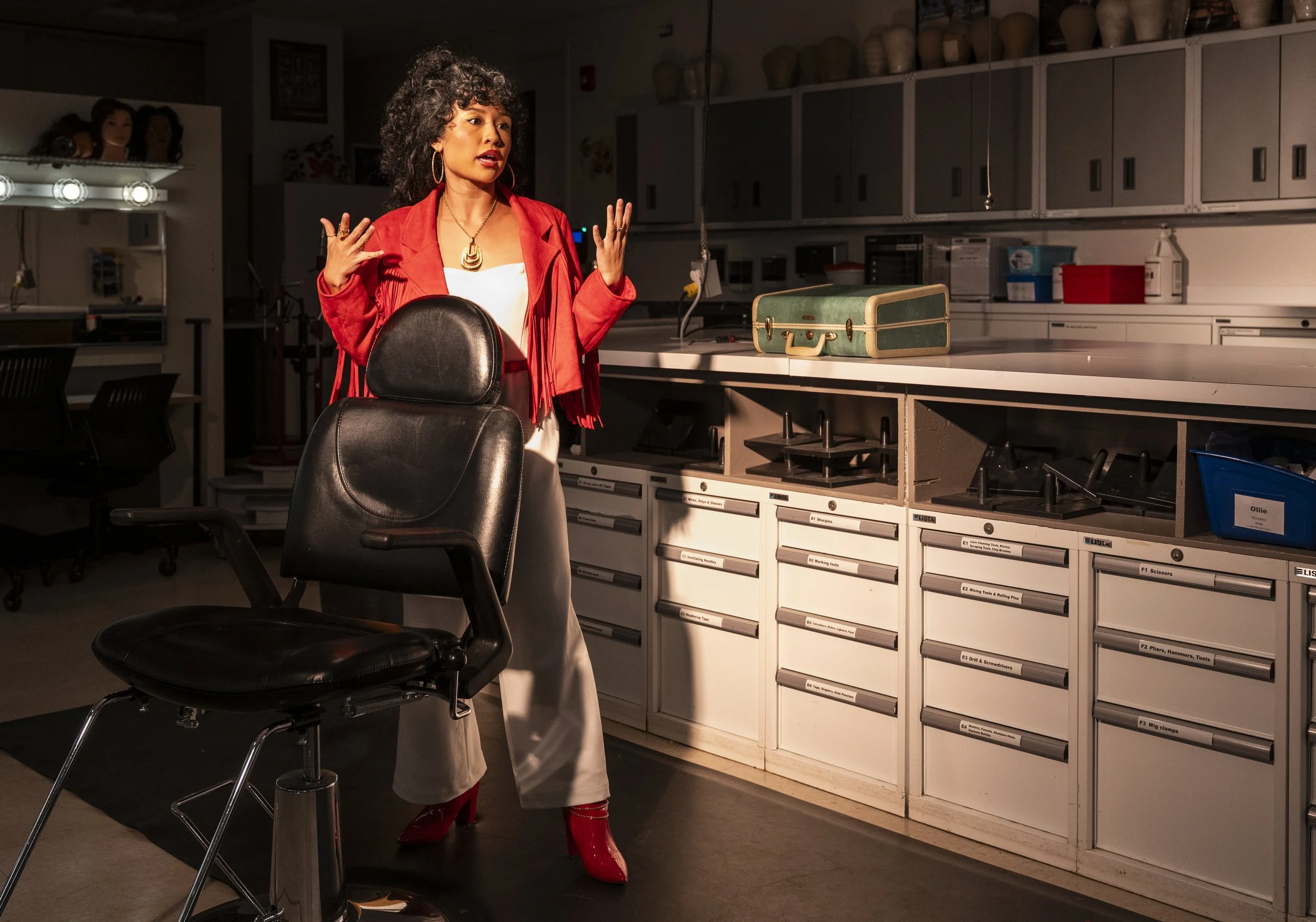 A woman with curly hair, wearing a red jacket, white top, and beige pants, stands in a laboratory or workshop, gesturing with her hands near a black chair and counter with drawers and storage compartments.