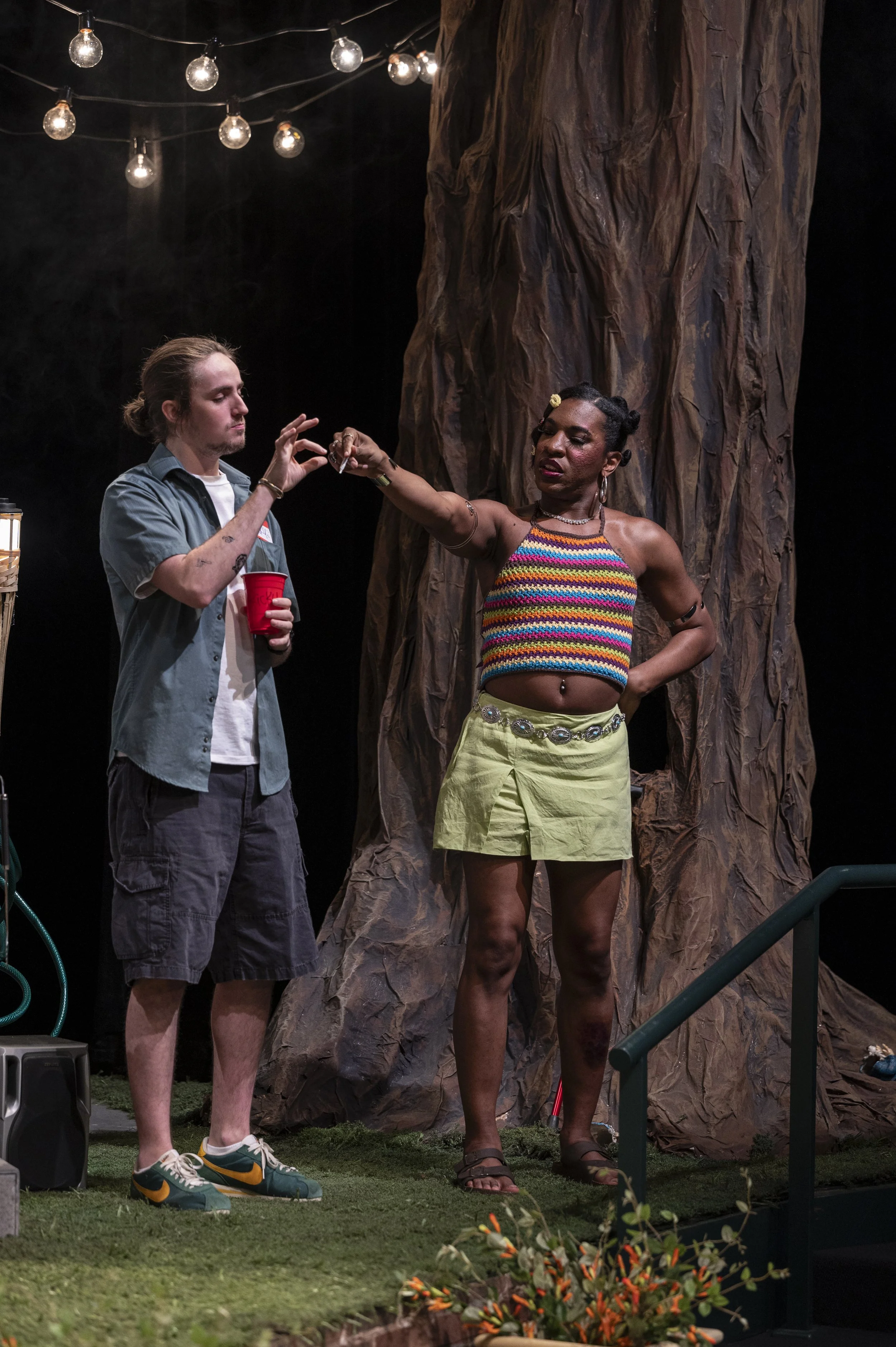 Two people are on stage, one woman with a colorful striped top and light green skirt, and a man in casual clothing holding a red cup. The woman is reaching out to the man, with a large tree-like structure behind them and string lights overhead.