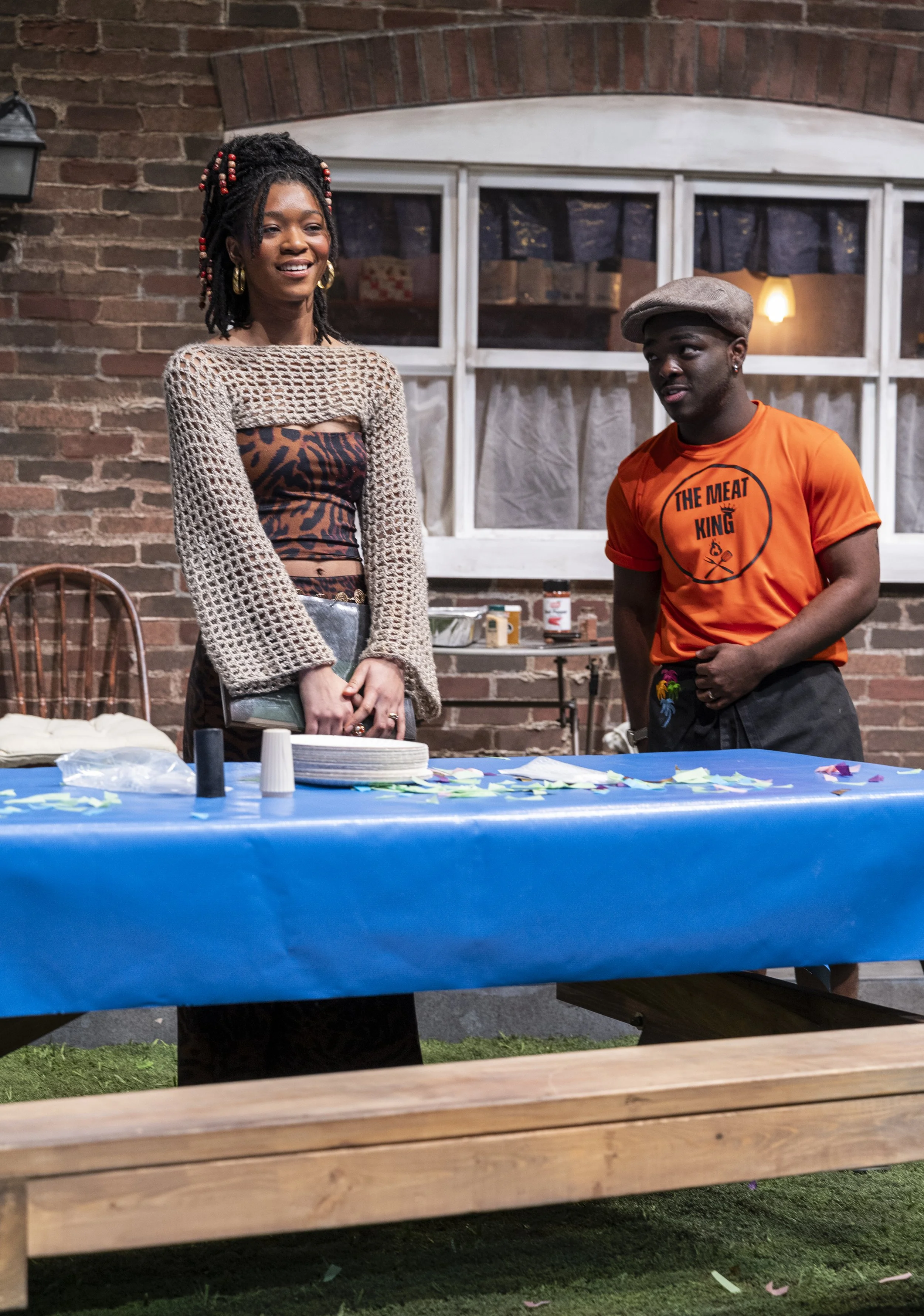 A woman with dreadlocks and a man wearing an orange T-shirt and a flat cap stand behind a table covered with a blue tablecloth. The woman is smiling, and the man is looking at her. There are paper plates, cups, and scattered paper items on the table.