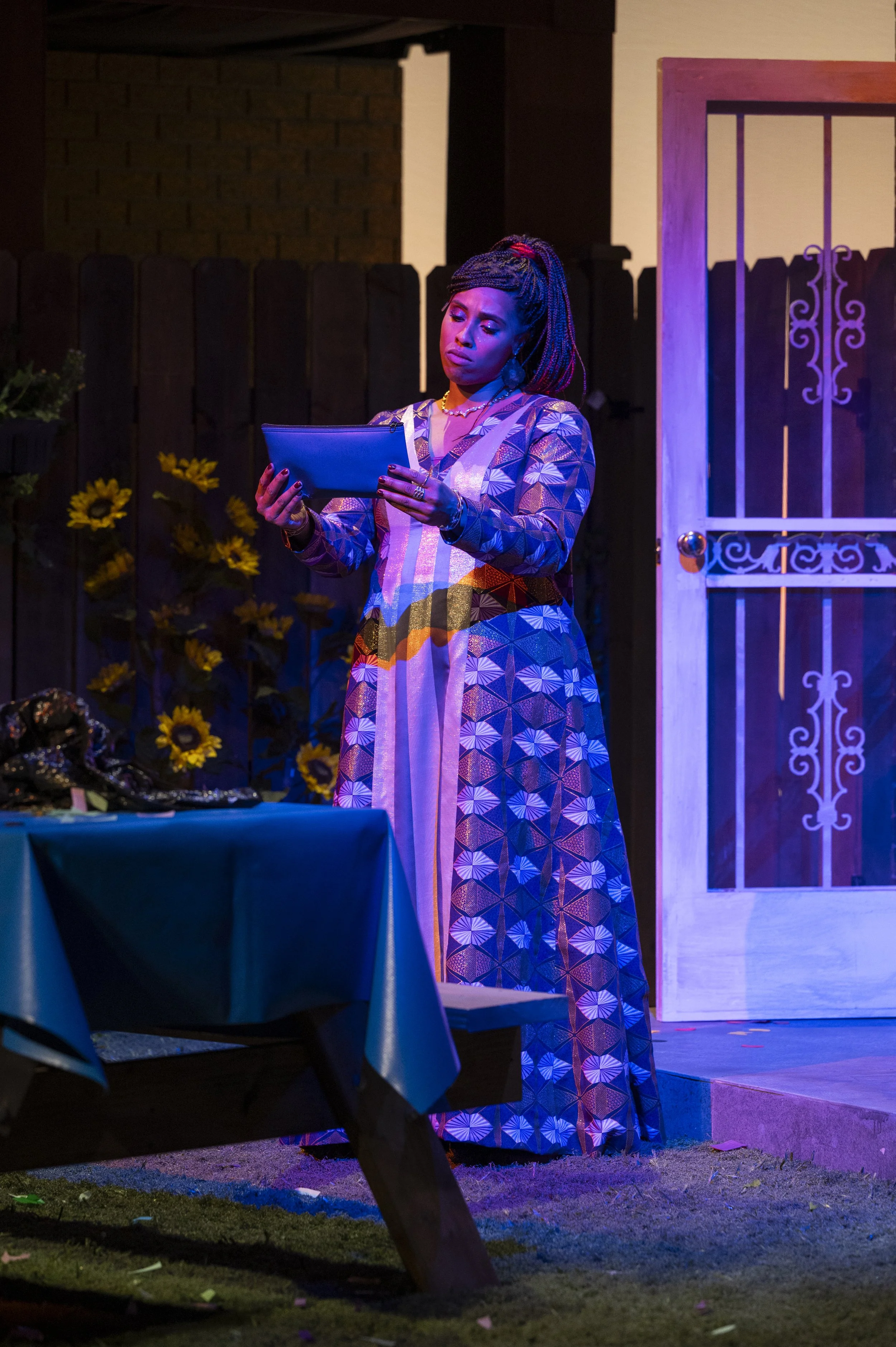 A woman in a colorful patterned dress standing on stage, reading from a tablet, with a wooden fence, sunflowers, and a white door in the background.