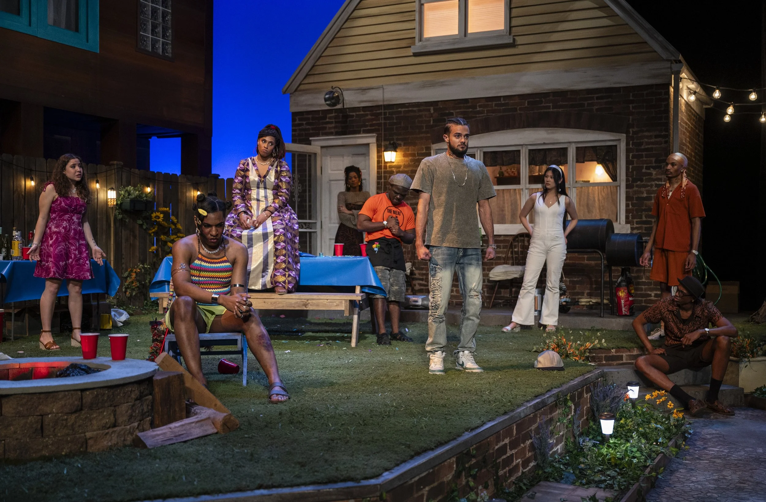 People gathered outdoors at night in a backyard, some sitting and some standing, with a house and string lights in the background, during a social event or party.