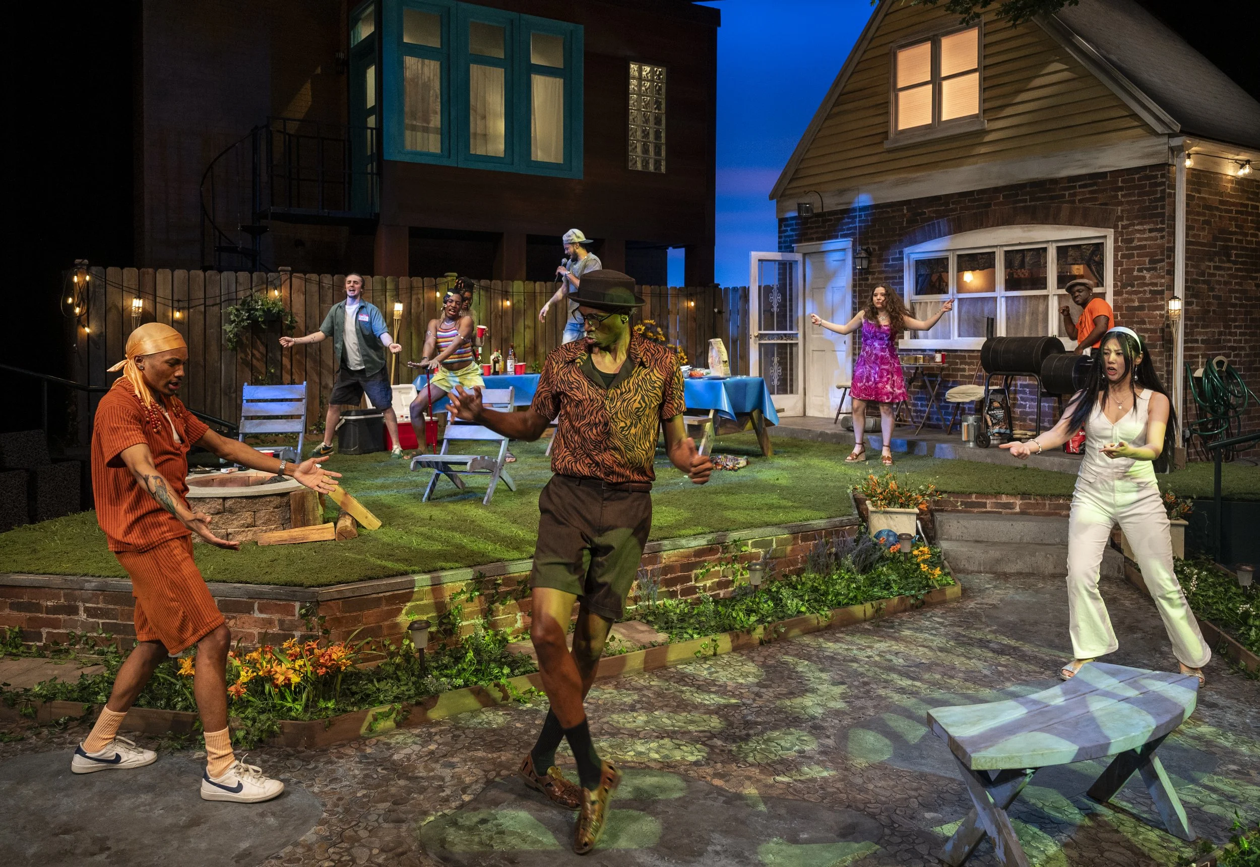 A lively backyard party scene with multiple people dancing and socializing. In the foreground, a woman in white is dancing, and a man in a patterned shirt and hat is dancing with outstretched arms. To the left, a woman in orange is facing the man, wi
