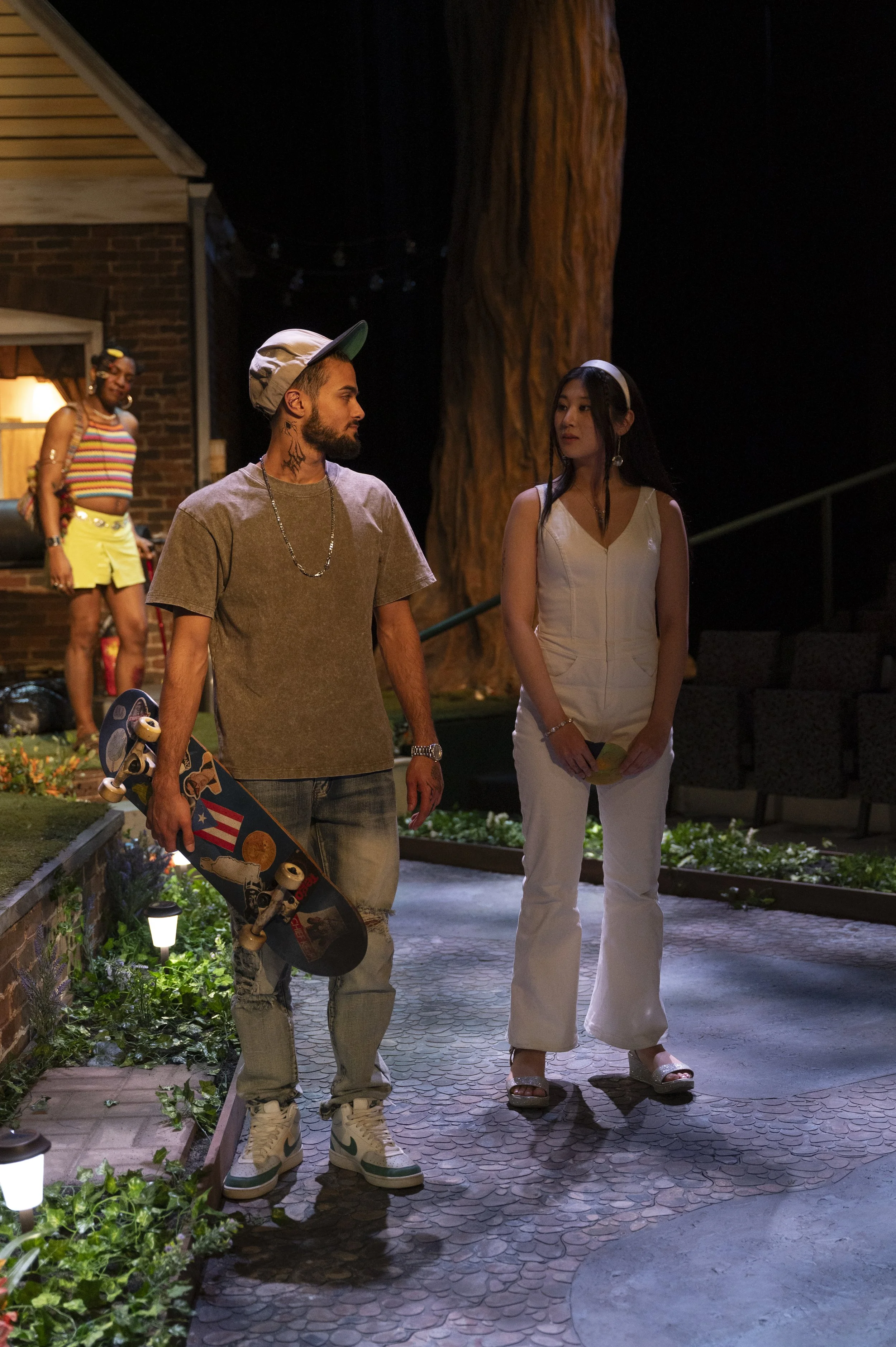 Two people are walking outside at night, the man with tattoos holding a skateboard and wearing sneakers, the woman dressed in white pants and top, holding a yellow fan, with a person in colorful attire in the background near a house.