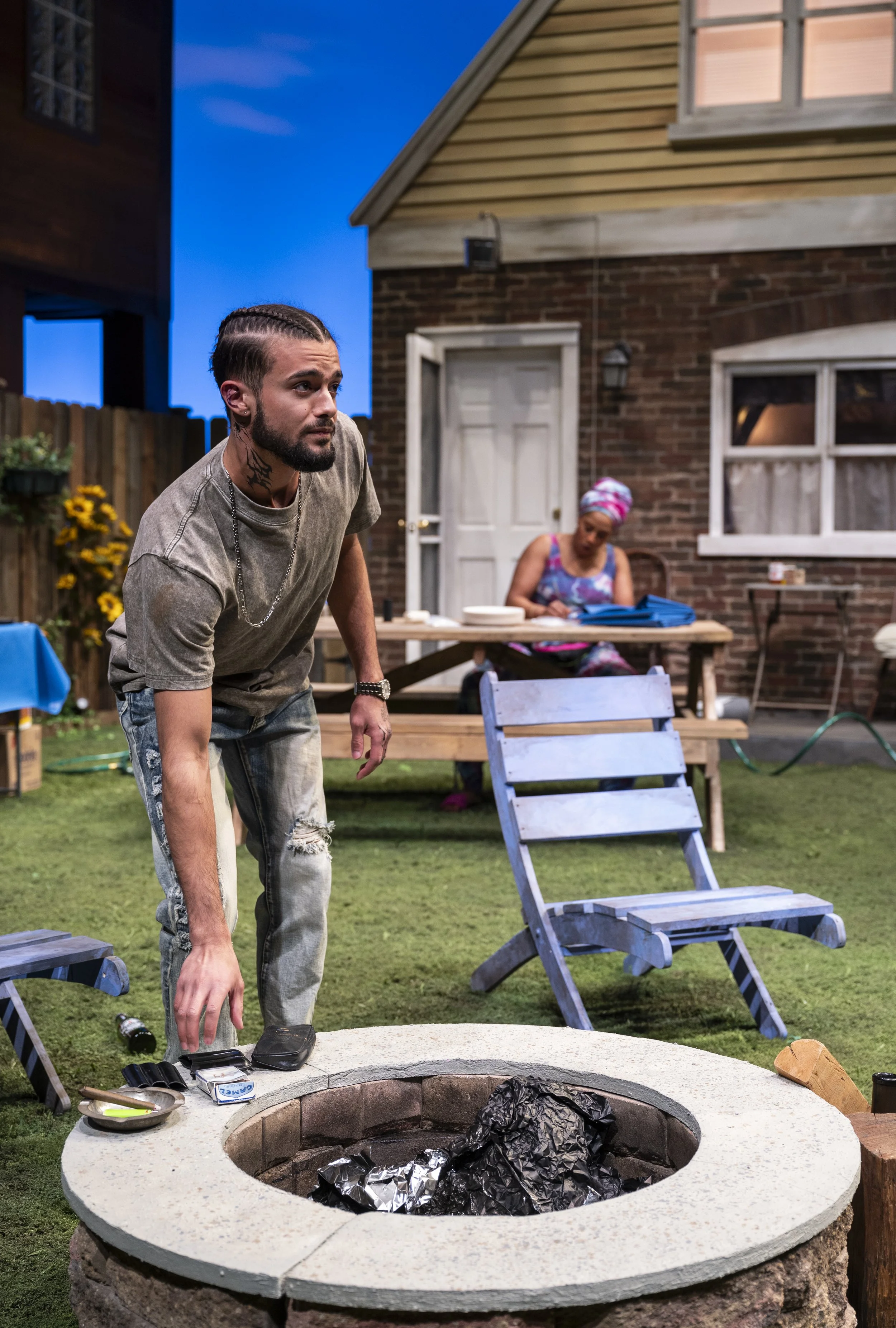 Man with braided hair and tattoos leaning over a firepit in a backyard during evening, with woman in colorful dress and headscarf sitting at a table in the background.