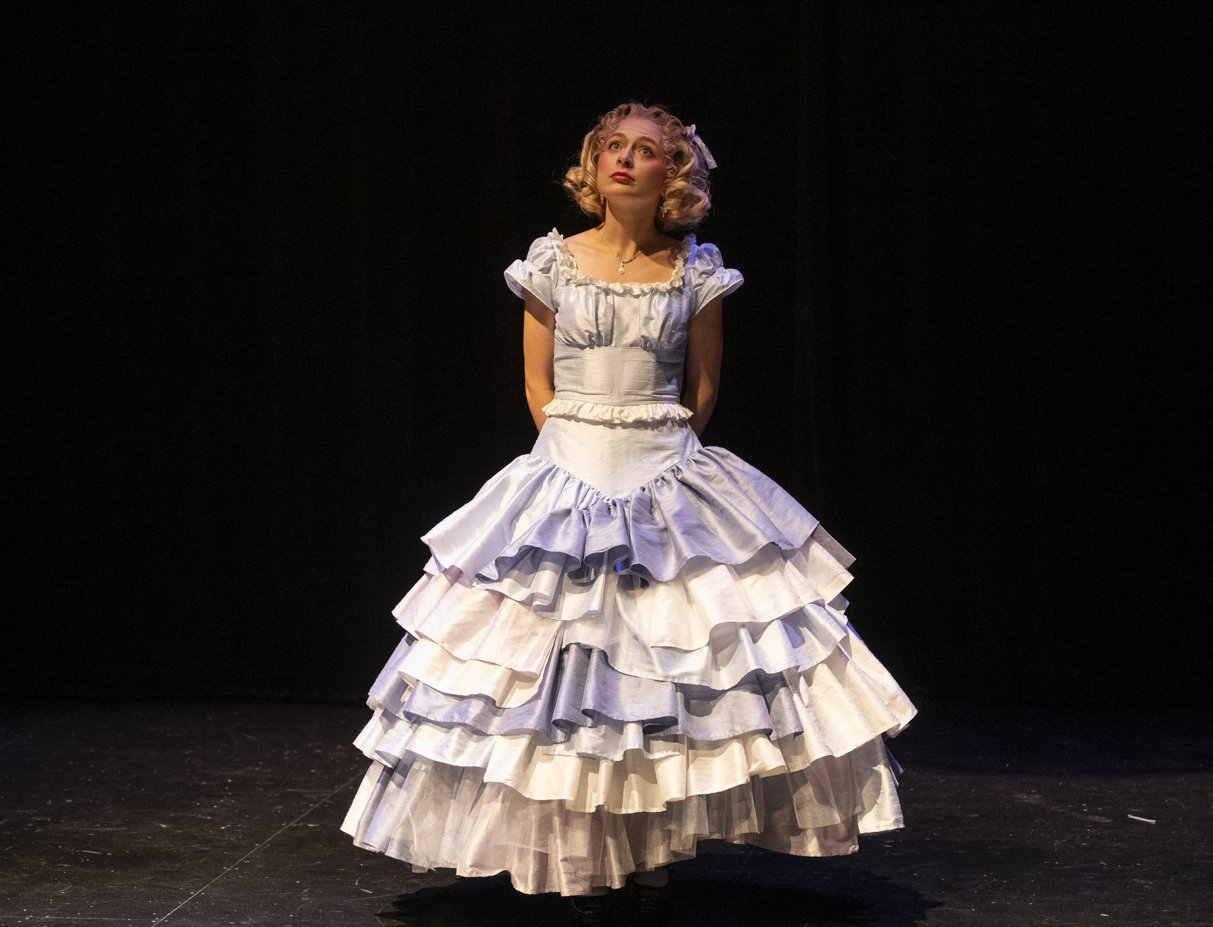 A woman in a vintage white, purple, and cream layered dress standing on a dark stage, looking upward.