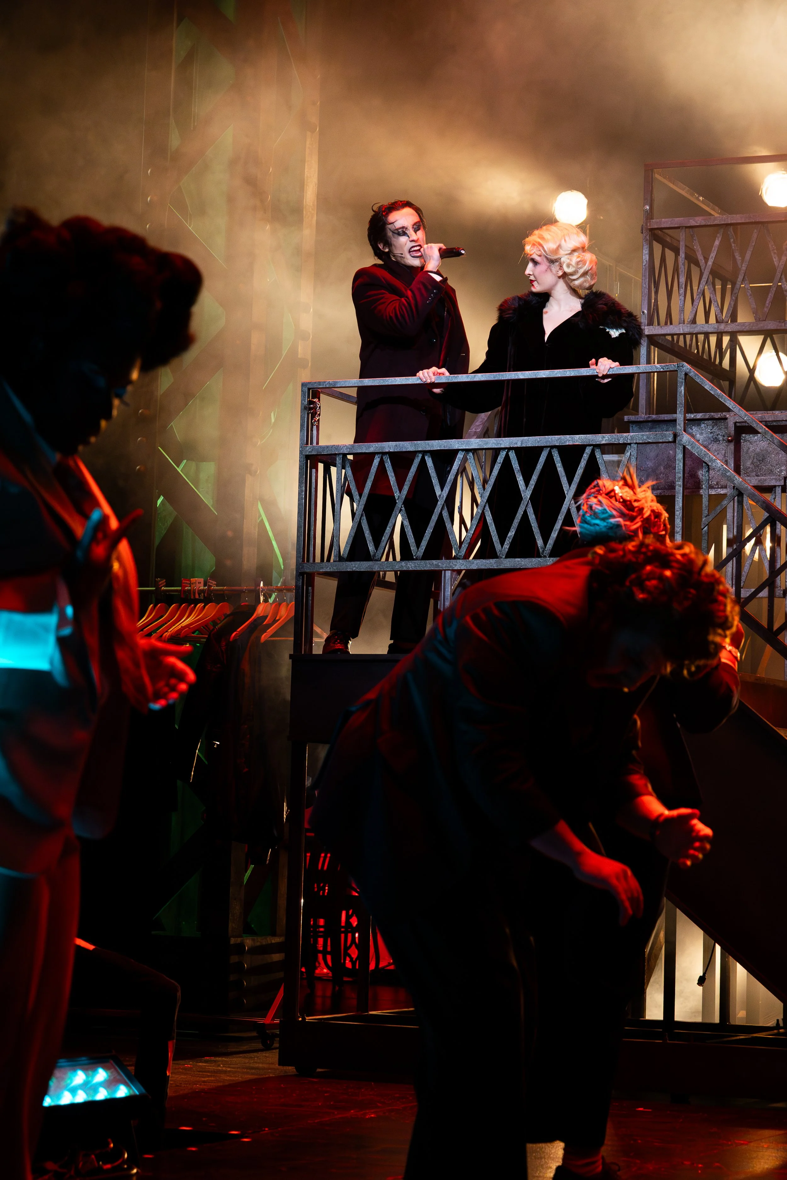 Scene from a theatrical performance, with a singer on an upper platform holding a microphone, a woman beside them, and individuals below stage with one using a smartphone and another bent over in a pose, set against smoky, dramatic lighting.