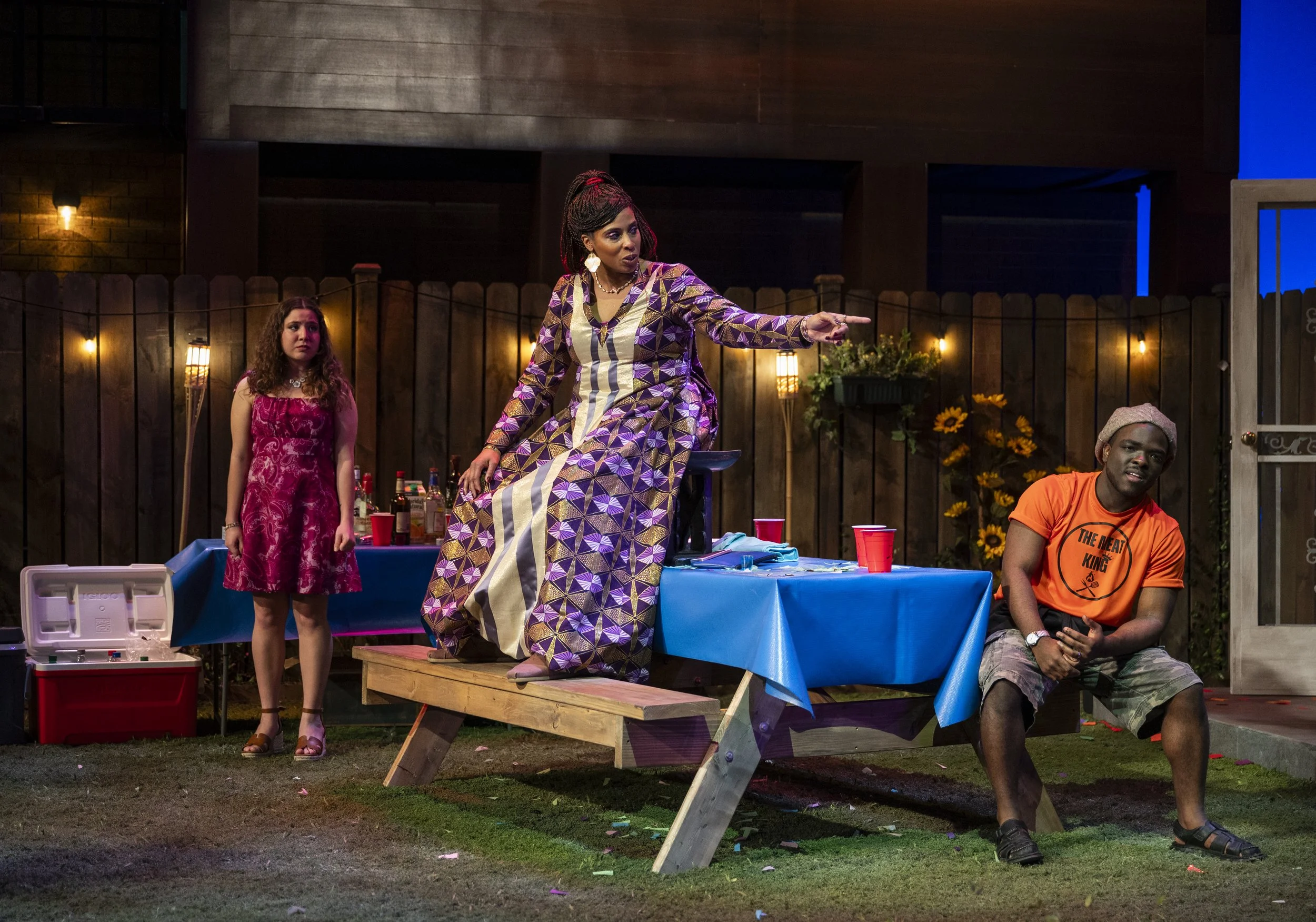Three people at a backyard party; woman in a colorful patterned dress is sitting on a picnic table and pointing, young woman in a pink dress is standing behind, man in an orange t-shirt and shorts is sitting on the ground. There are red cups and a co