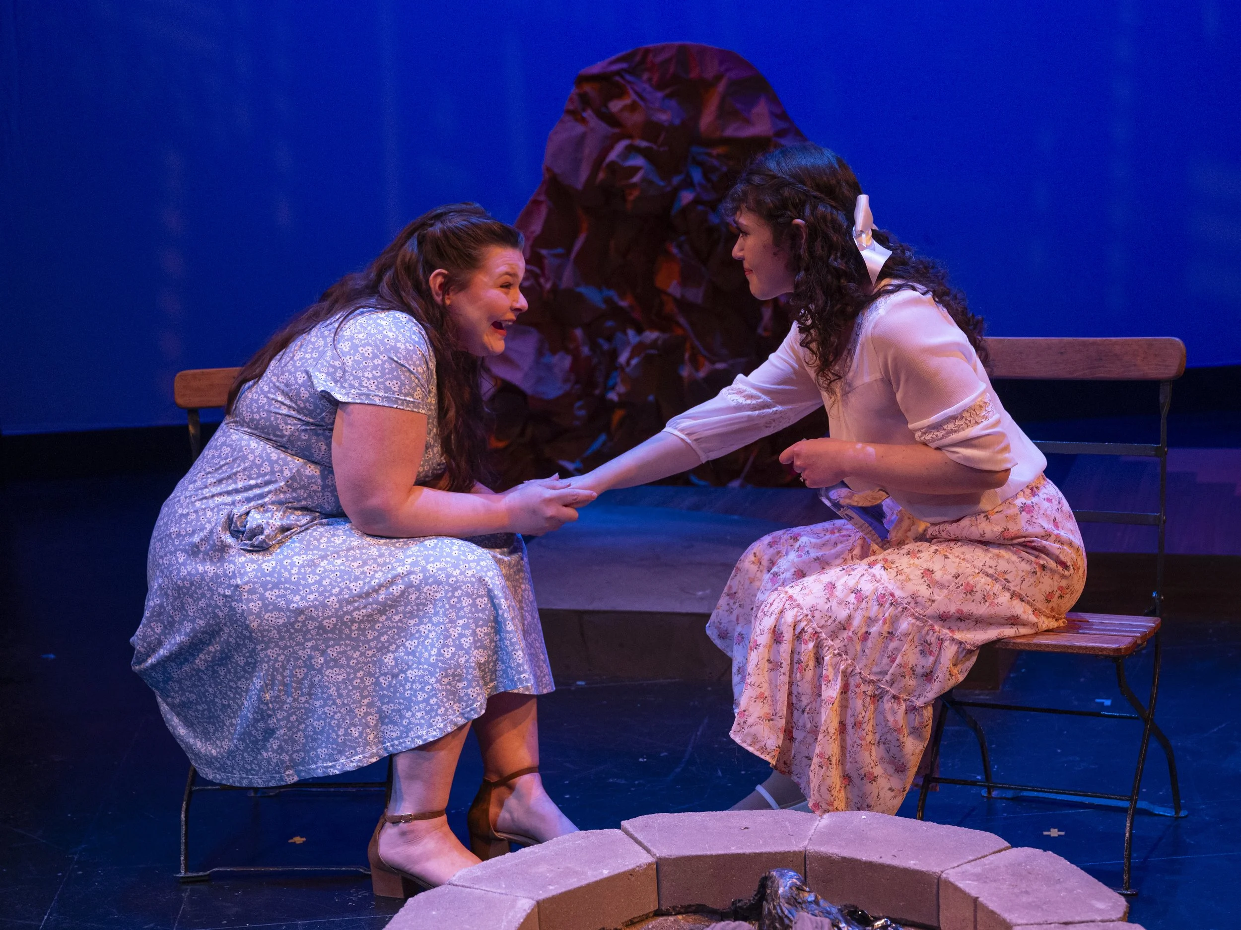 Two women sitting on benches facing each other, holding hands, performing a scene in a play. One woman is smiling and laughing, wearing a light blue floral dress with heels, and the other is in a white top and pink floral skirt with a white bow in he