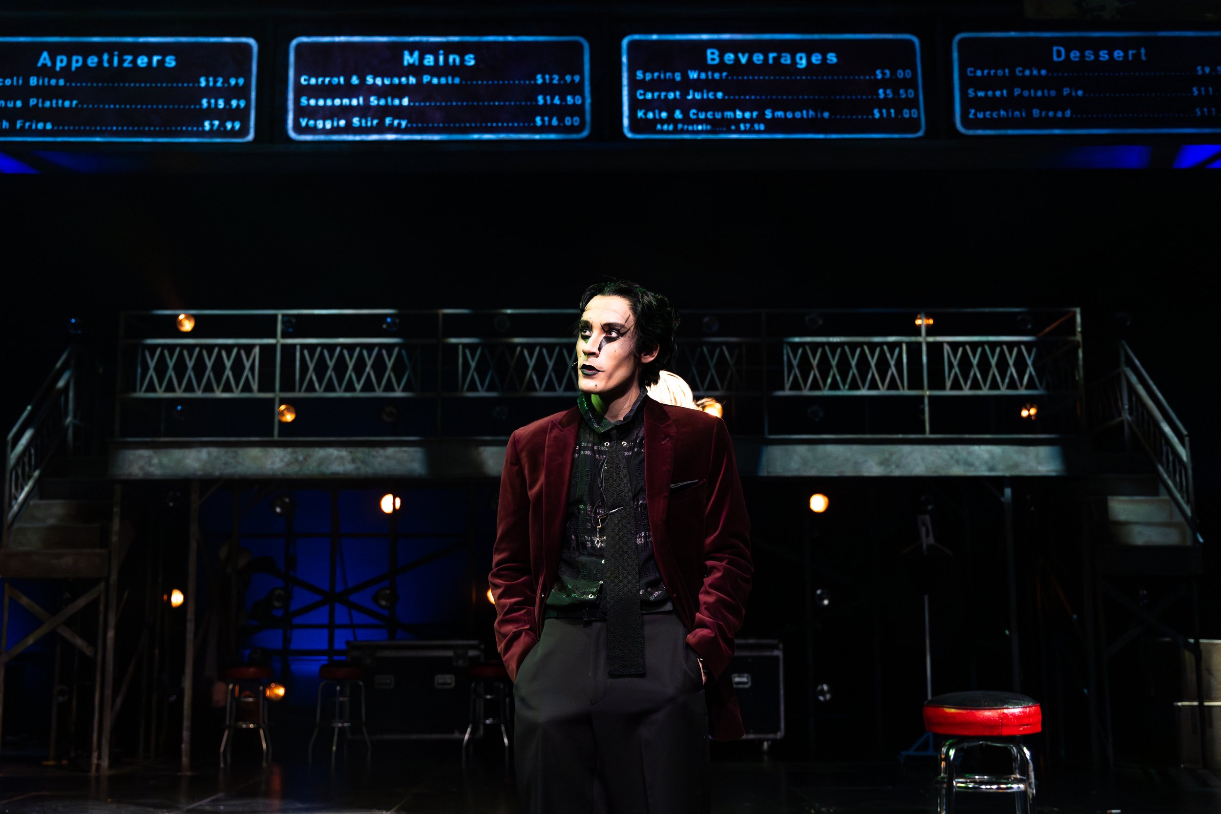 A man dressed in a dark red velvet blazer and black pants, with styled dark hair and makeup, stands on a dimly lit stage with a dark background, and a large menu board illuminated above him.