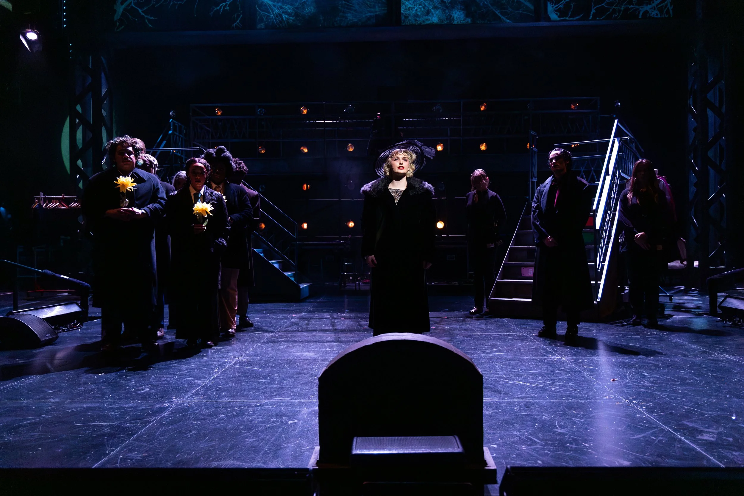 'Black the Broadway musical cast performing on stage with one woman standing in the center wearing a black coat and hat, others hold yellow flowers, on a dark theater stage with stairs and lights in the background.
