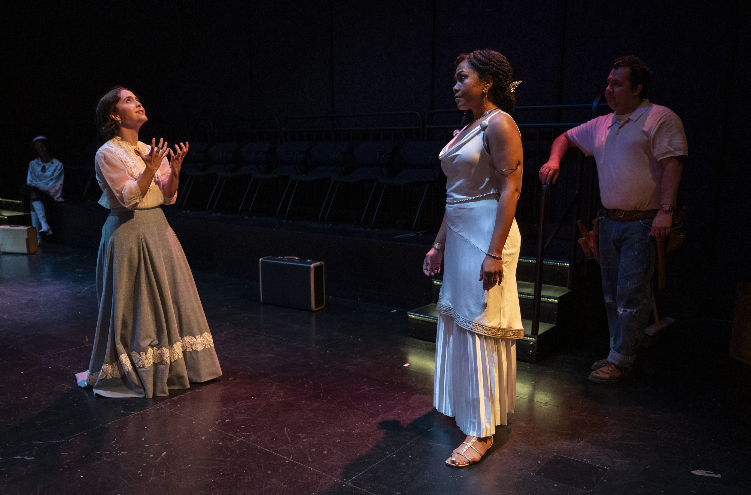Three women and one man on a stage, with two women in vintage style dresses and the man in casual clothing, engaged in a dramatic scene during a theatrical performance.